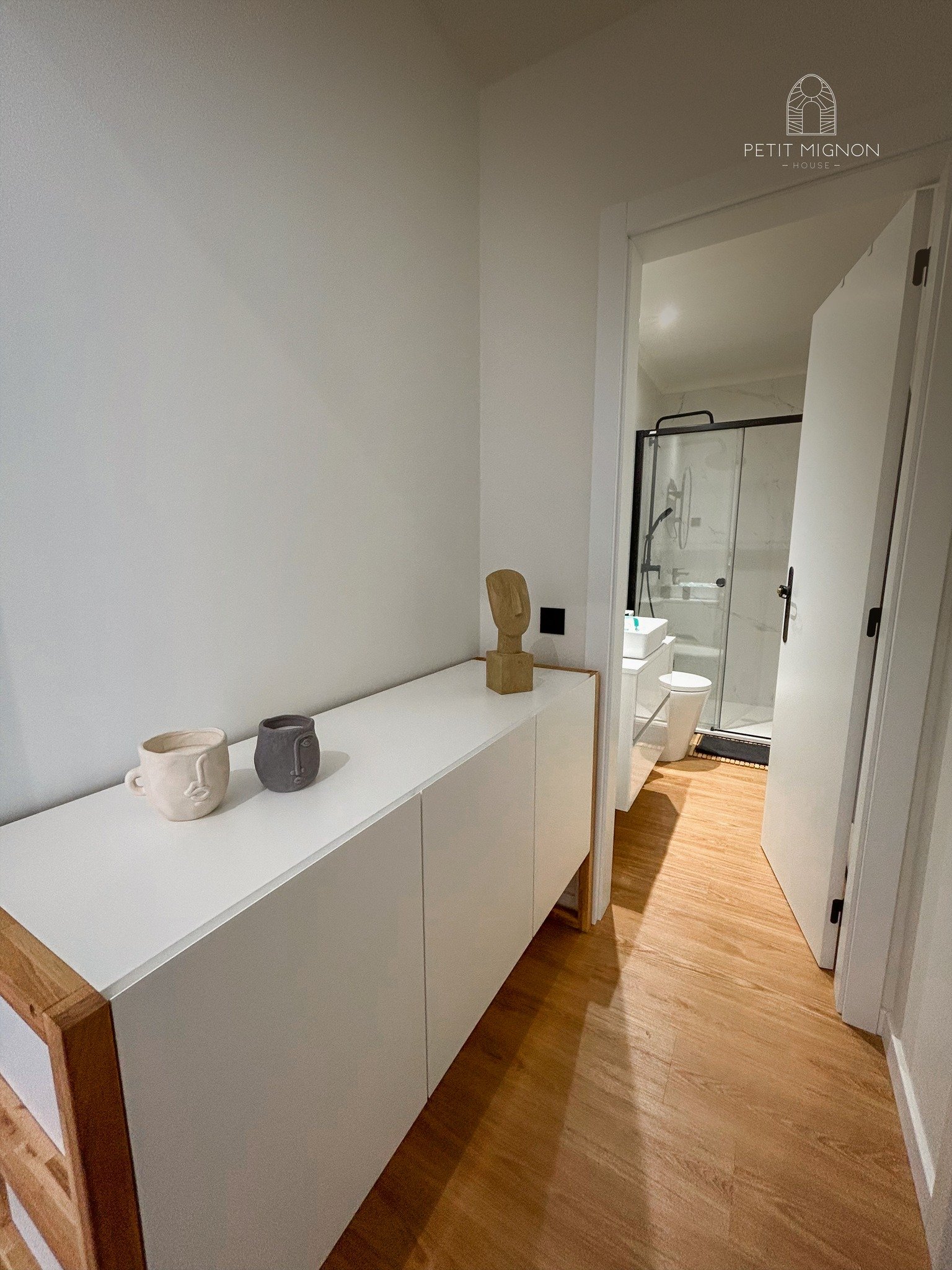 Corredor de um apartamento com um móvel branco decorado com três vasos de cerâmica e uma escultura. Ao fundo, banheiro com box de vidro, pia branca, vaso sanitário e piso de madeira.