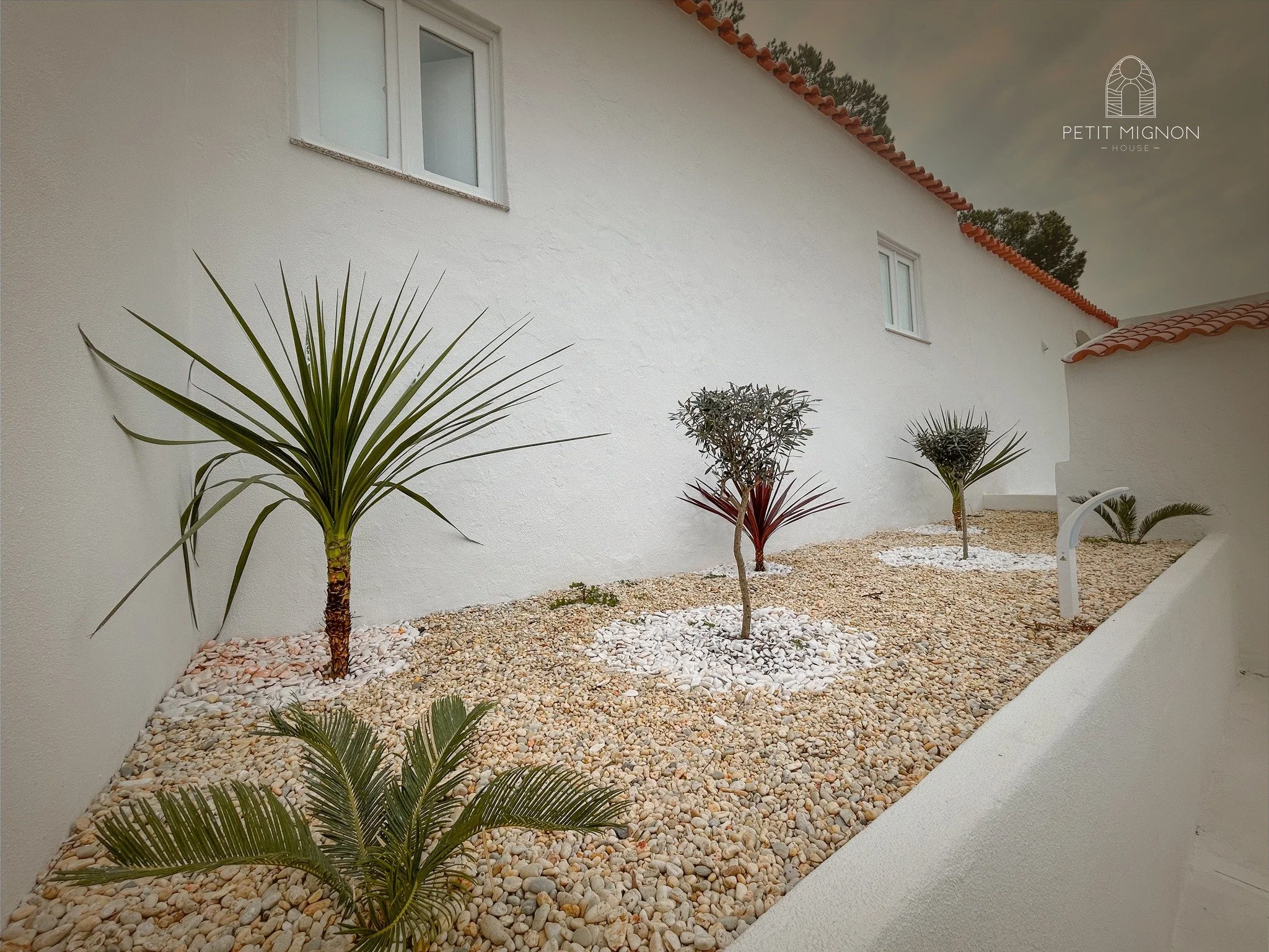 Jardim com pedras, árvores e plantas decorativas ao lado de uma casa branca com janelas e telhado de telhas vermelhas.