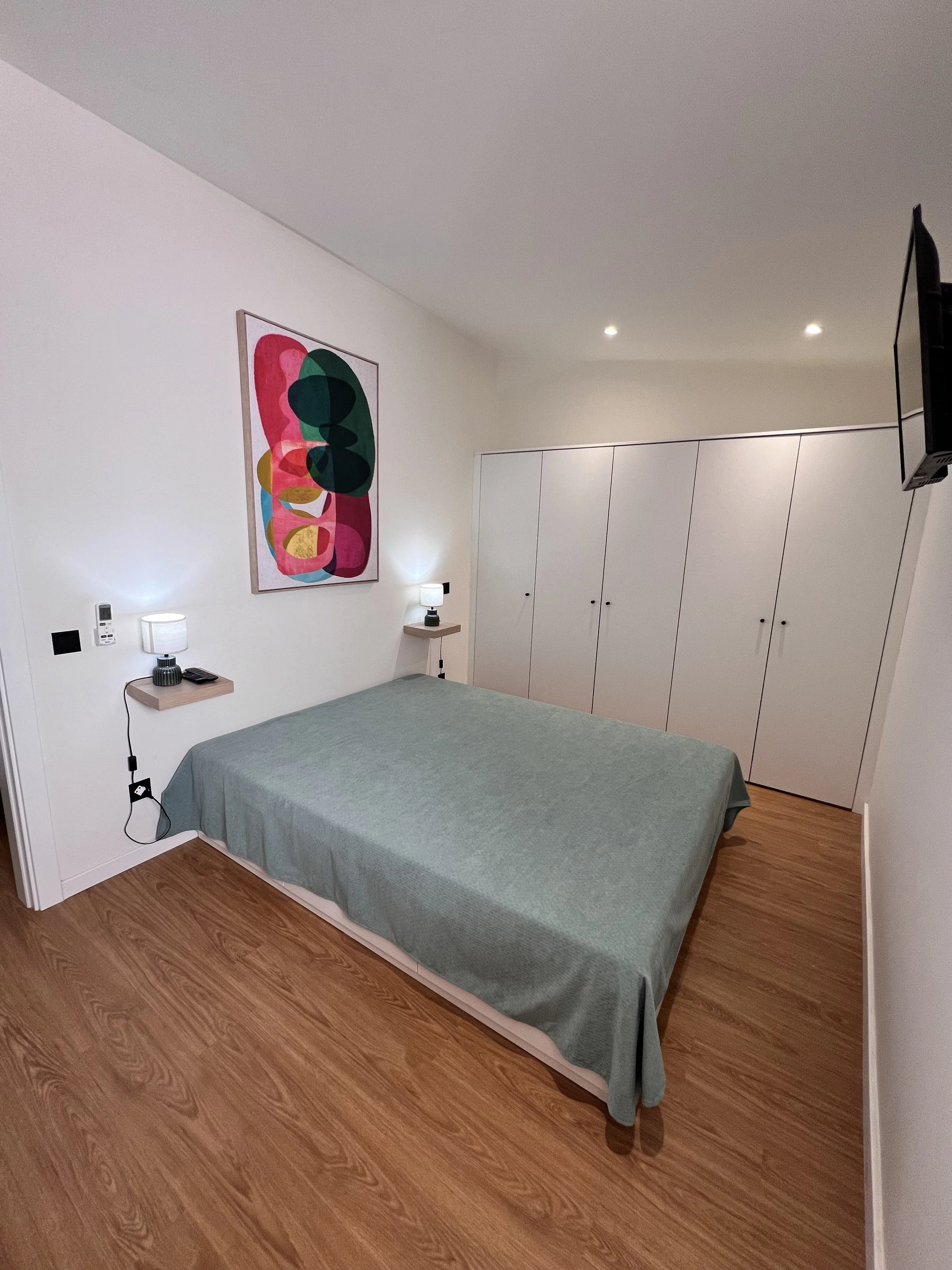 Quarto com cama de cabeceira baixa, roupa de cama verde, prateleiras ao lado com luminárias, armários brancos em uma parede, parede com quadro de arte abstrata colorida, televisão na parede, piso de madeira.