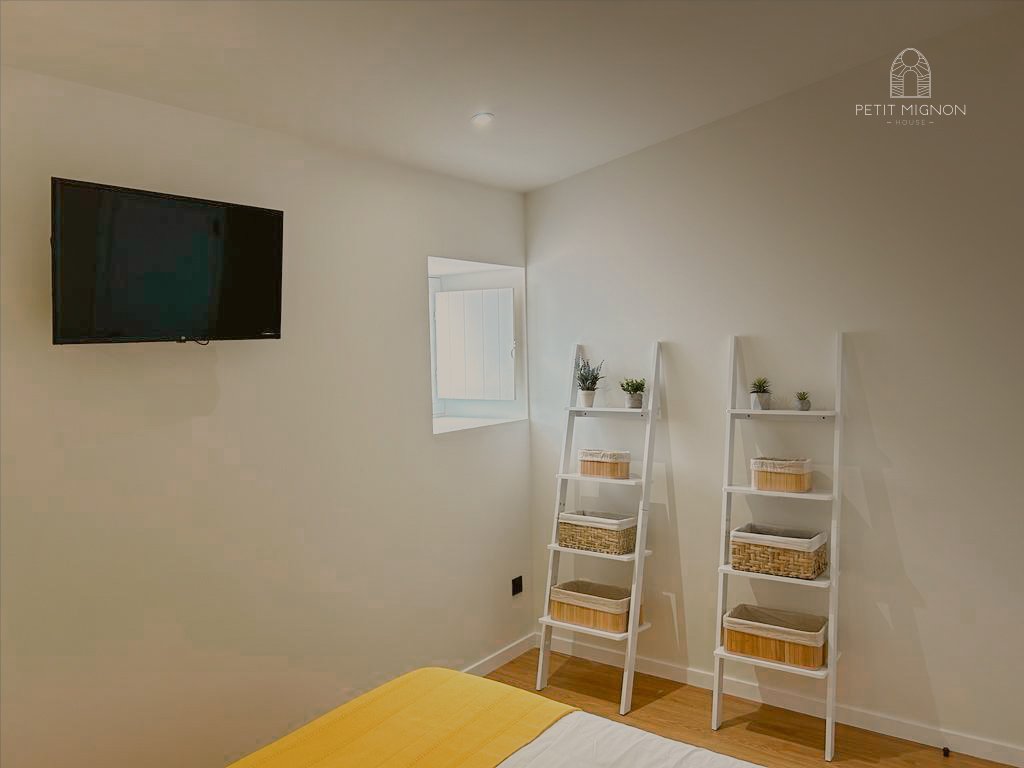Quarto com televisão na parede, cama com lençol amarelo, duas prateleiras brancas apoiadas no chão com cestas de armazenamento, uma janela pequena na parede ao lado da televisão, decorado com plantas pequenas.