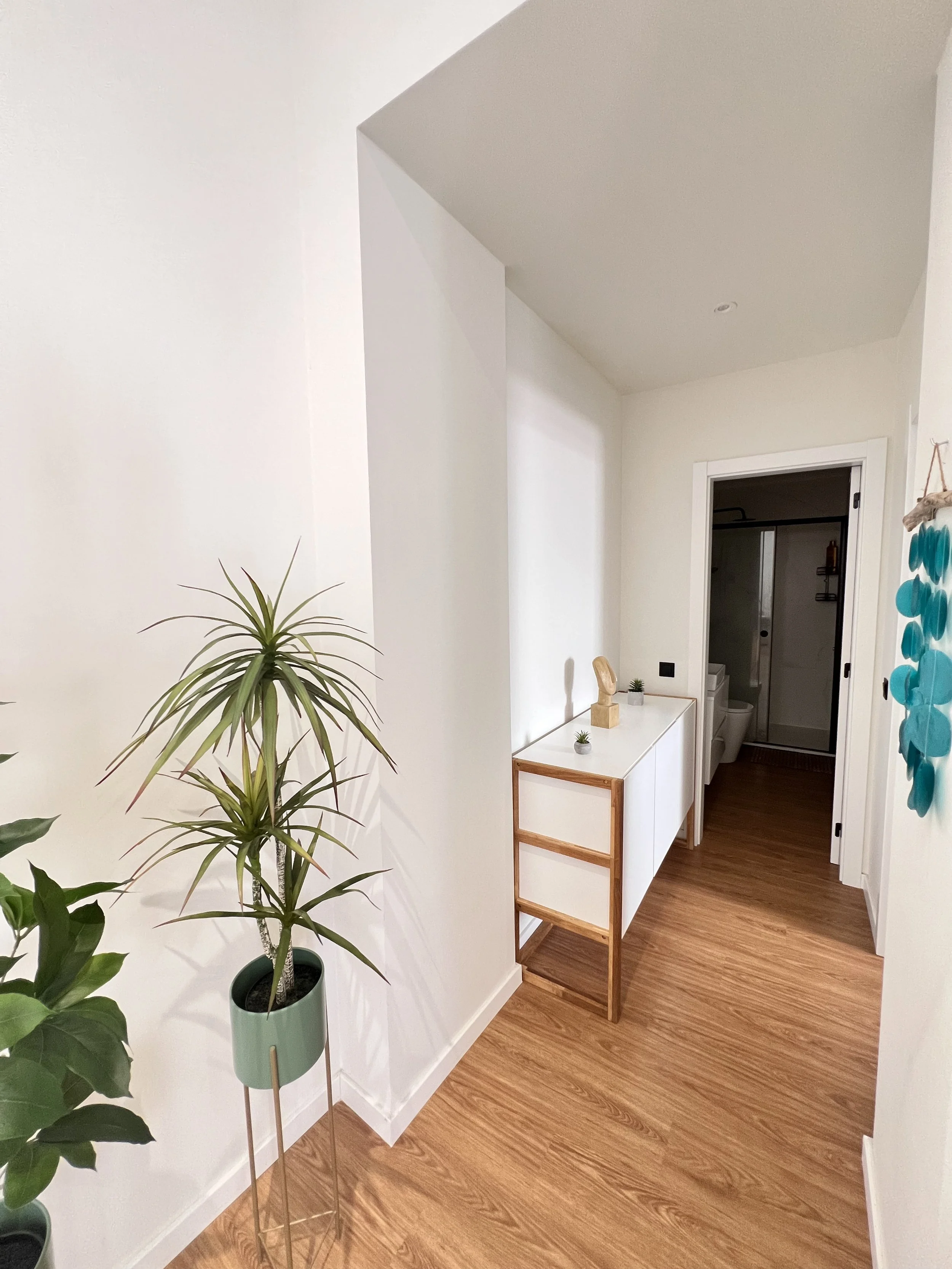 Corredor de casa com piso de madeira clara, paredes brancas, com plantas em vasos e móveis brancos e de madeira.