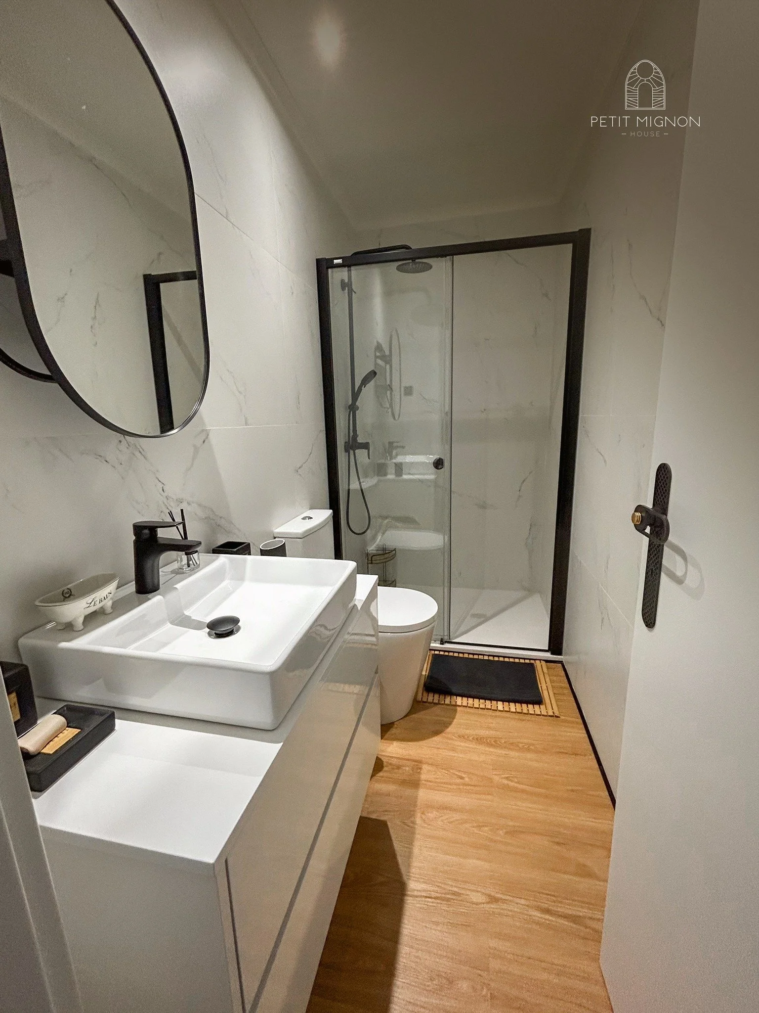 Banheiro com parede de mármore branco, pia com cuba retangular branca, espelho oval, vaso sanitário, box de vidro com porta de correr, piso de madeira e tapete preto.