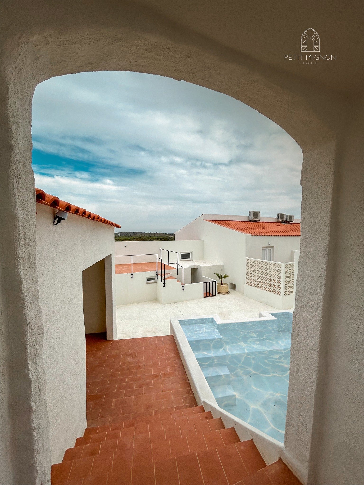 Vista de uma área de lazer com piscina, escadas de degraus, piso de azulejo vermelho, parede branca, teto de arco, céu nublado e algumas casas brancas com telhados vermelhos ao fundo.