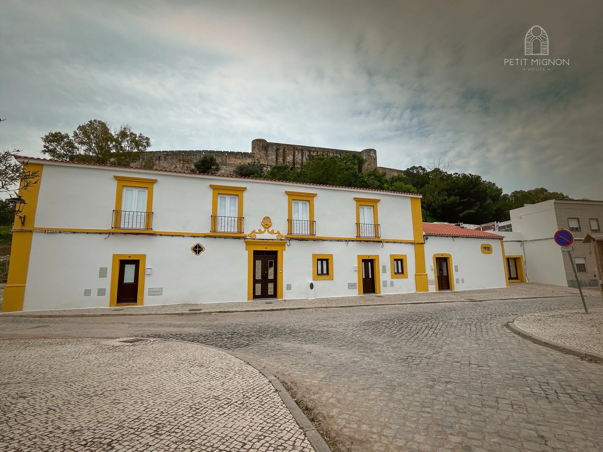 Casa branca com detalhes em amarelo na esquina de uma rua de paralelepípedos, ao fundo há uma colina com uma fortificação antiga e árvores.