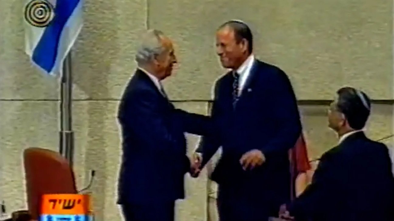 Two men in dark suits shaking hands at a ceremony, with a flag in the background and a seated individual observing.