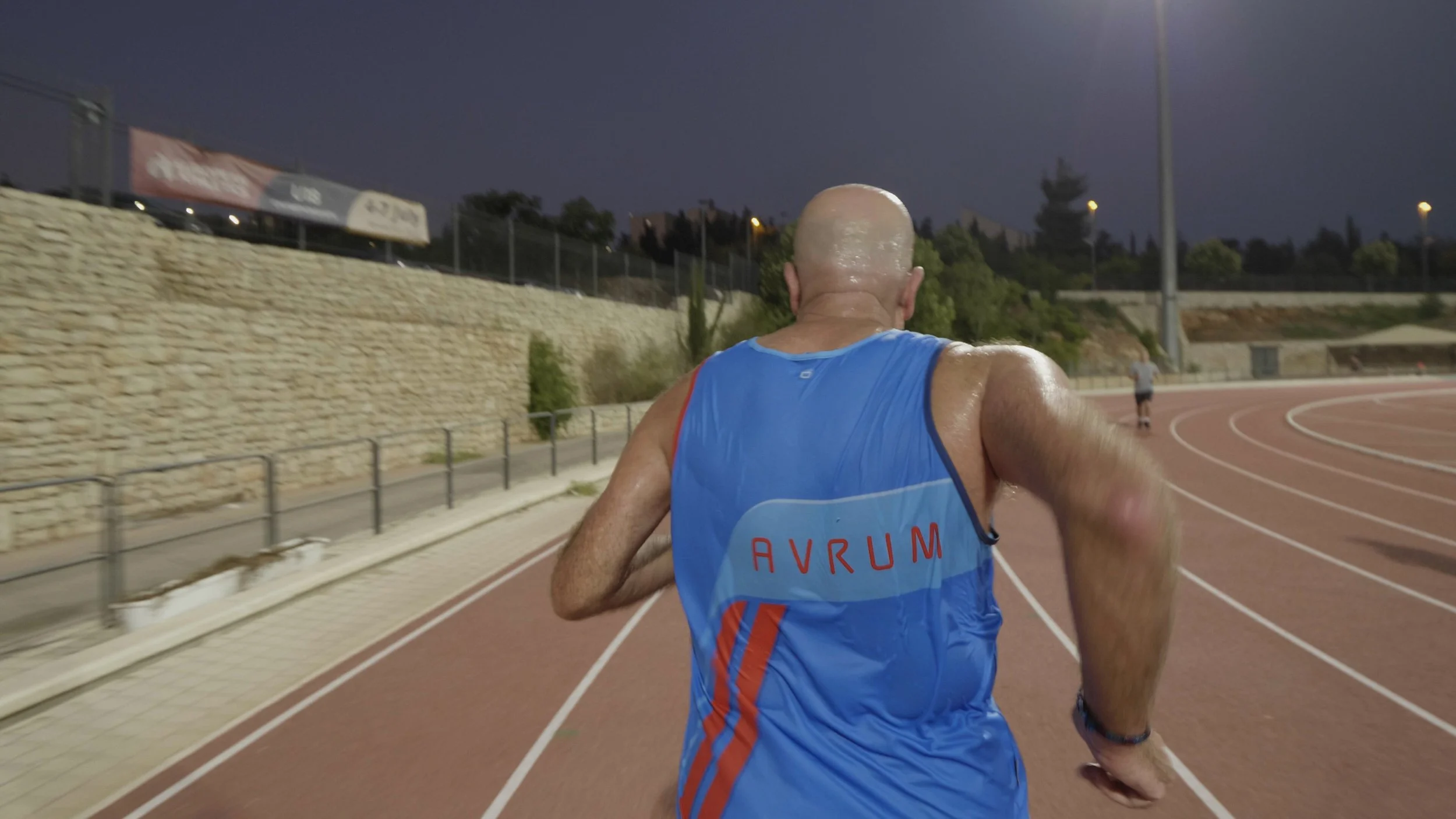 A male runner wearing a blue tank top with red and orange accents on a running track at dusk.