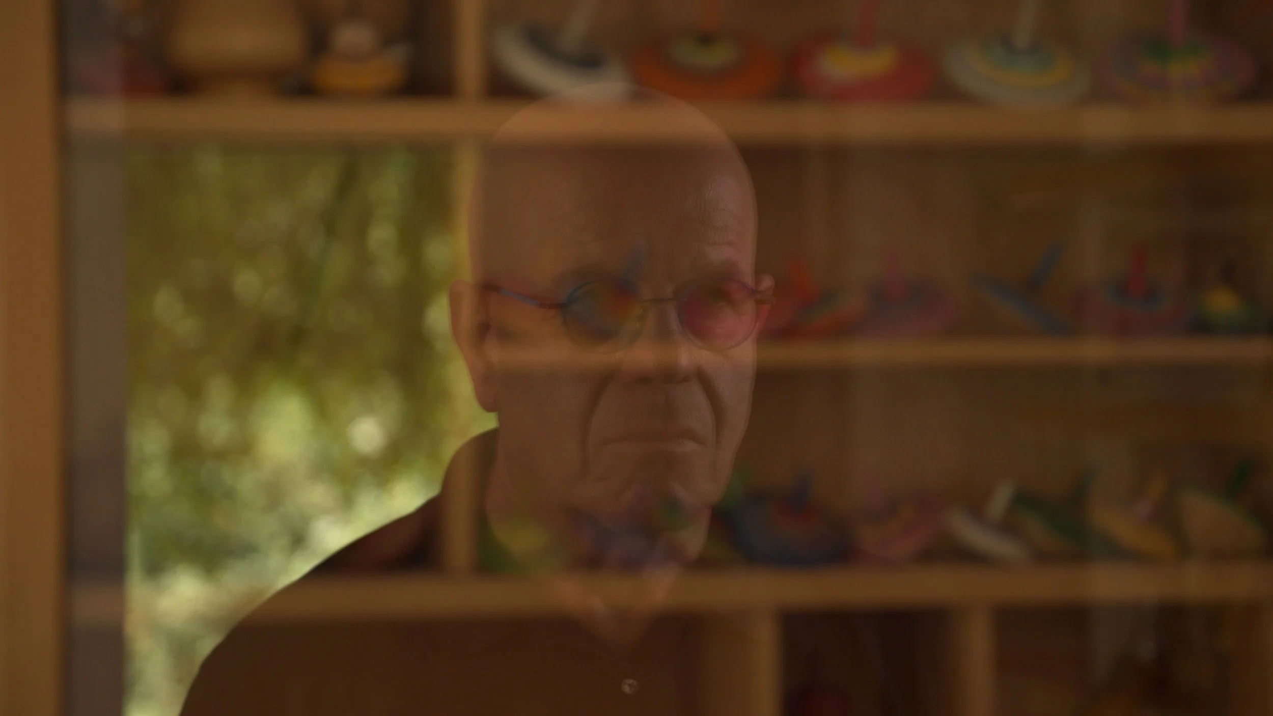 A reflection of an elderly man with glasses, viewed through a glass window, with a wooden shelf of colorful items in the background.