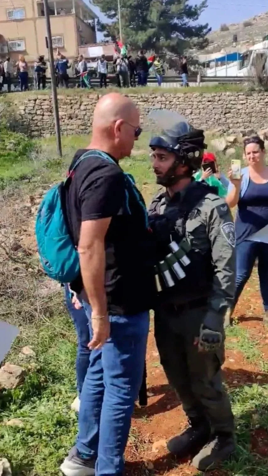 A bald man and a soldier face each other in a confrontation outdoors, with a group of people observing nearby and on a hill in the background.