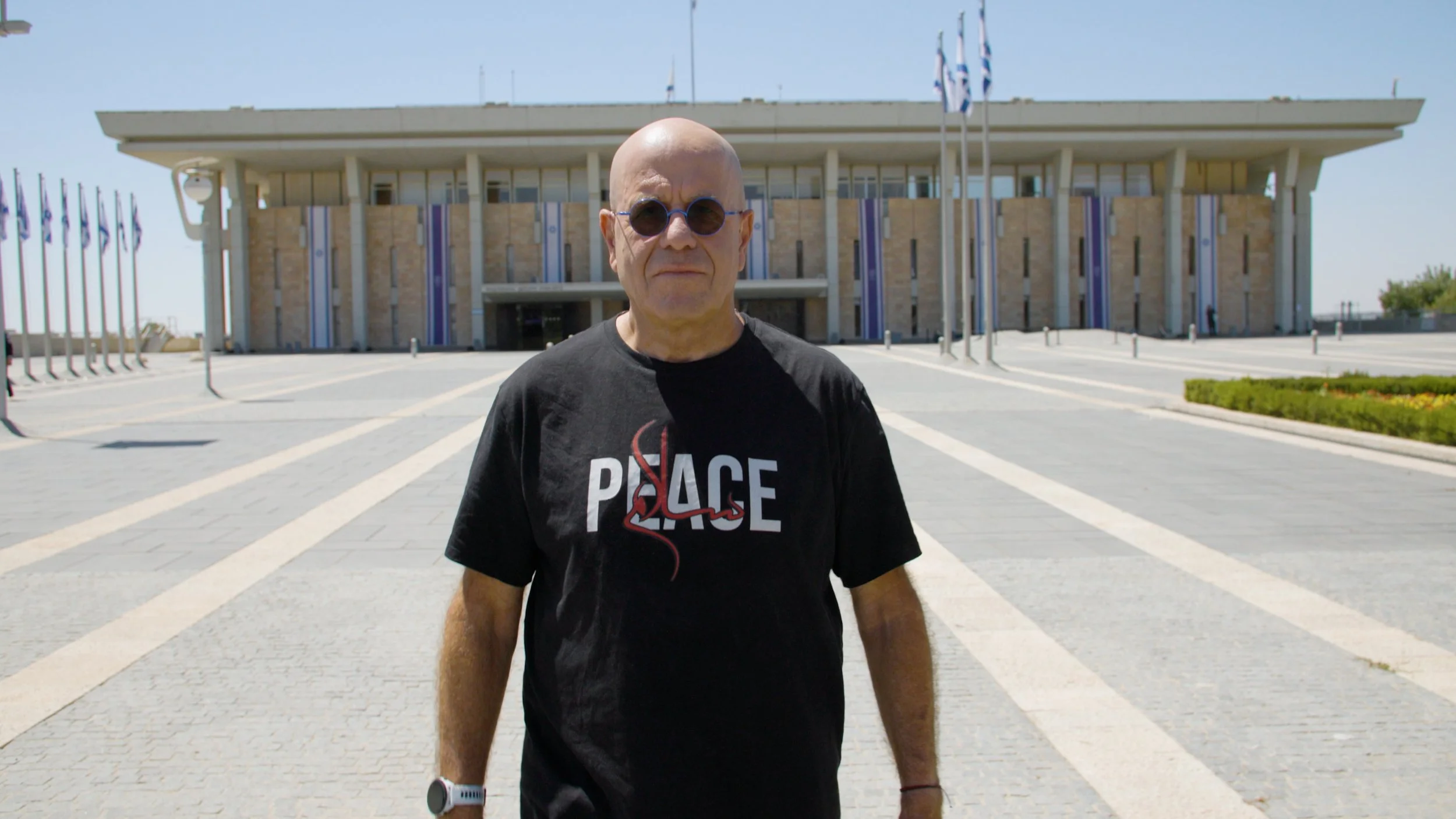 A man wearing sunglasses and a black t-shirt with the word 'PEACE' and a red ribbon stands outdoors on a paved walkway in front of the Knesset, a large modern building with multiple flagpoles and flags in the background.