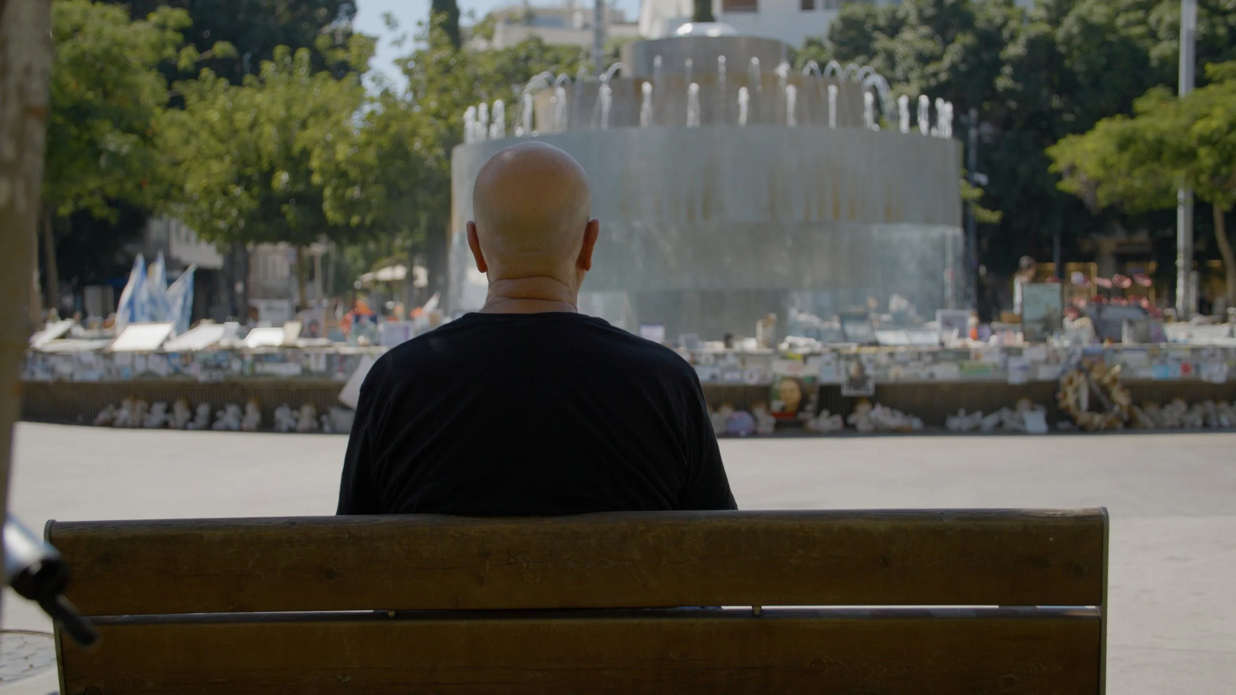 A bald man sitting on a wooden bench in front of a large fountain in a public square, with trees and buildings in the background.