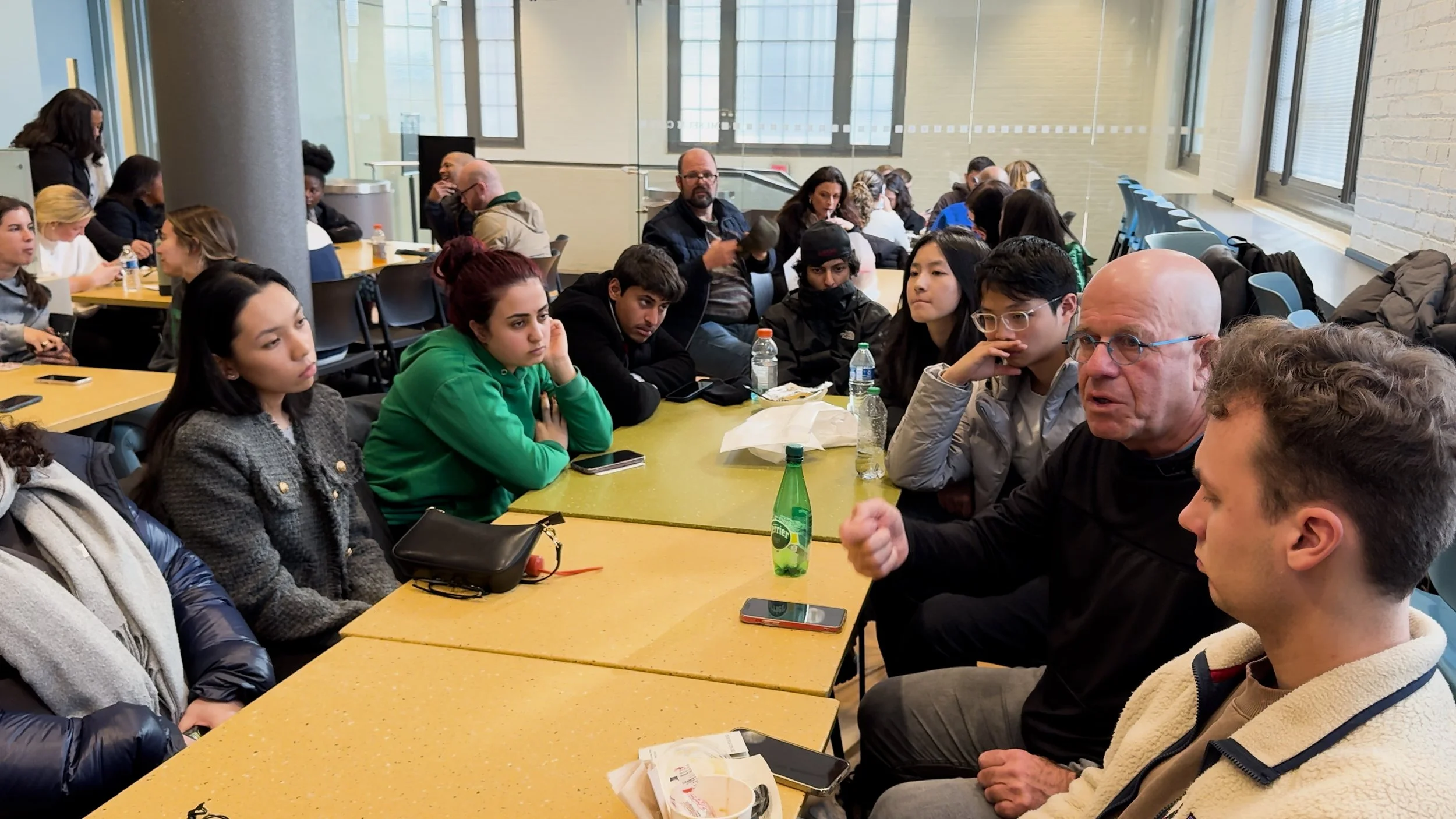 A group of diverse young people and older adults sitting around a yellow table engaged in a discussion or listening to a speaker, in a bright, modern room with large windows.