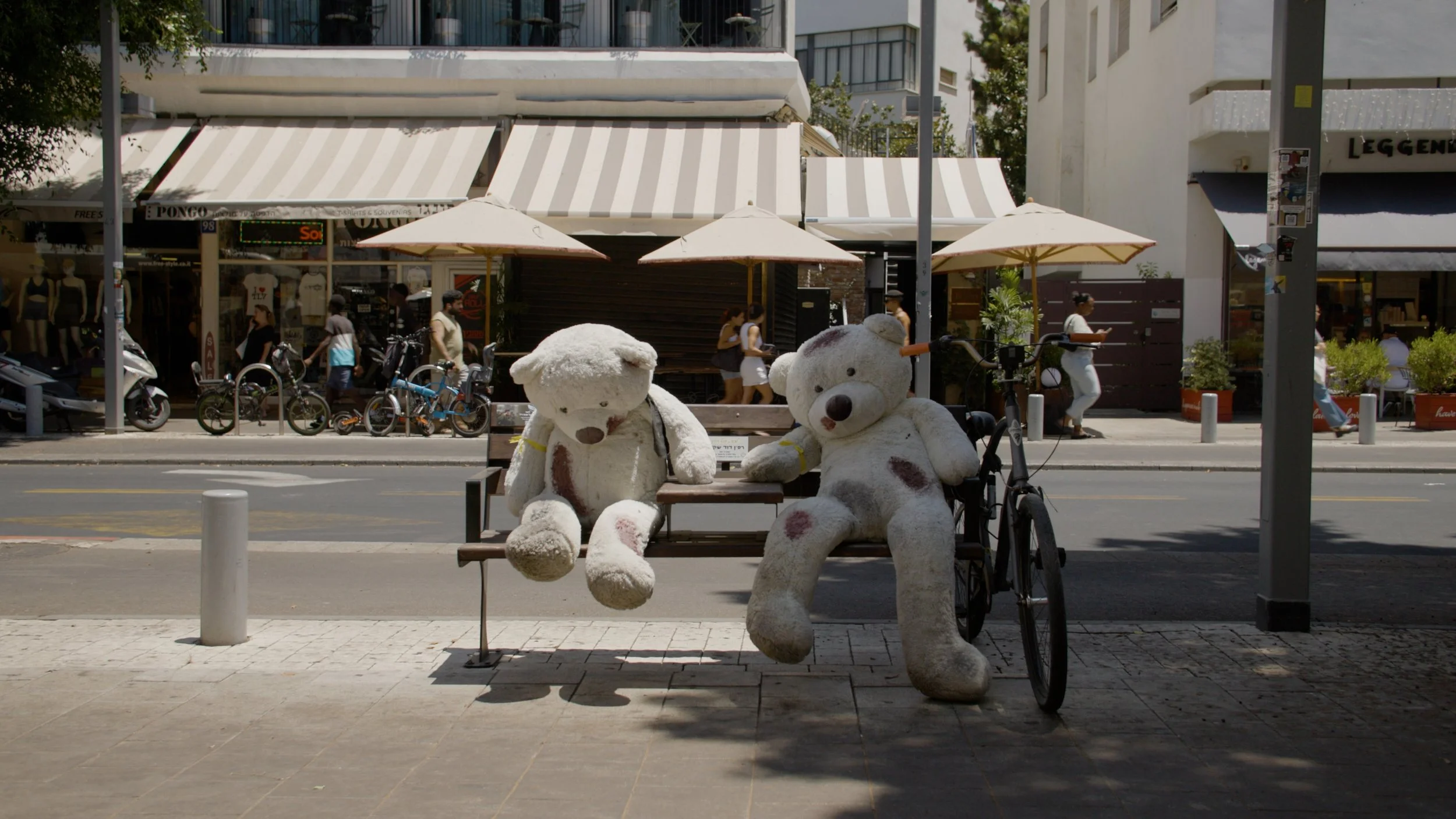 Two large blood stained teddy bears sitting on a park bench, on a sunny city street with shops and pedestrians in the background.