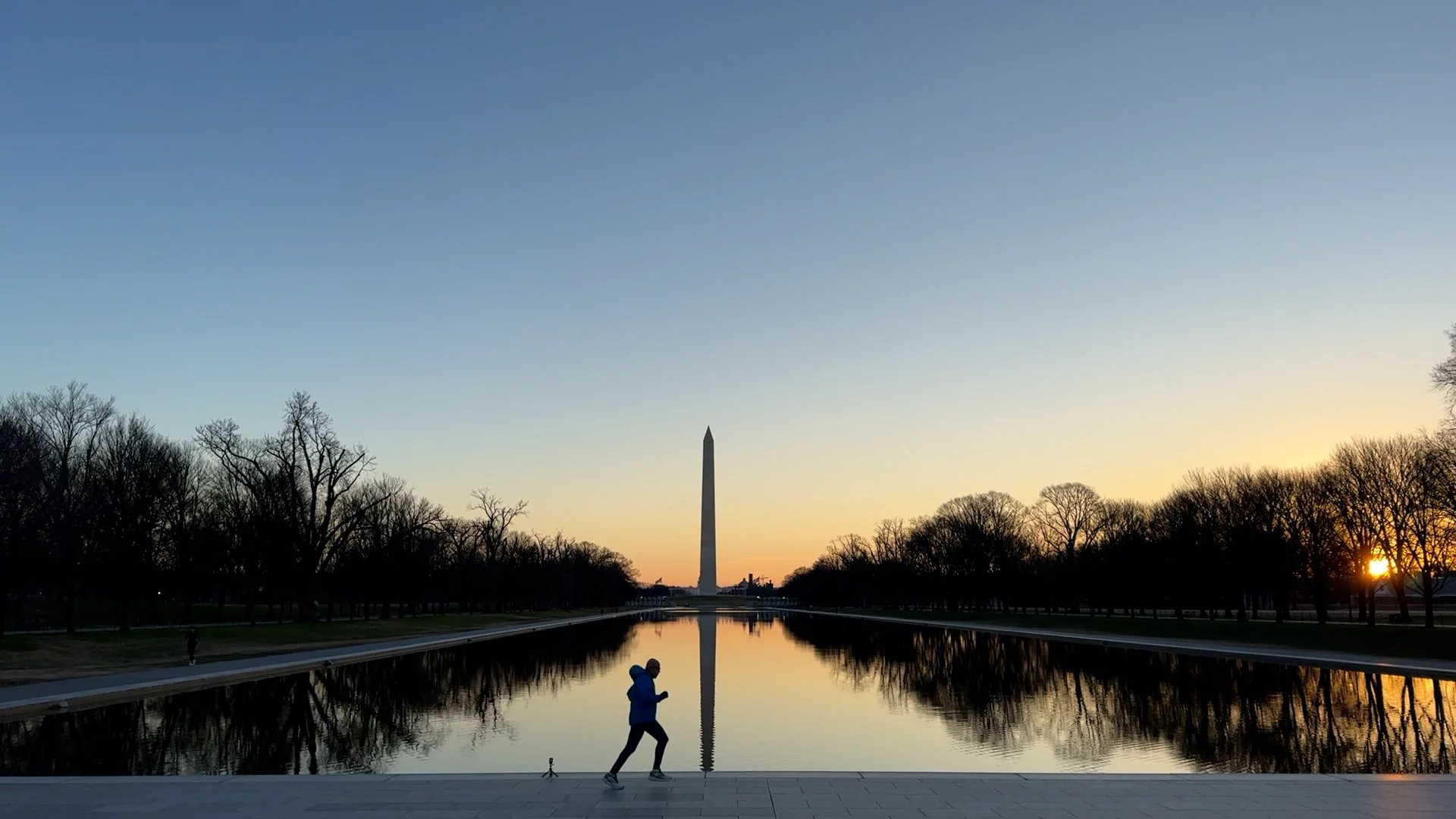 Silhouette of a person jogging in front of the Reflecting Pool with the Washington Monument in the background at sunrise or sunset.