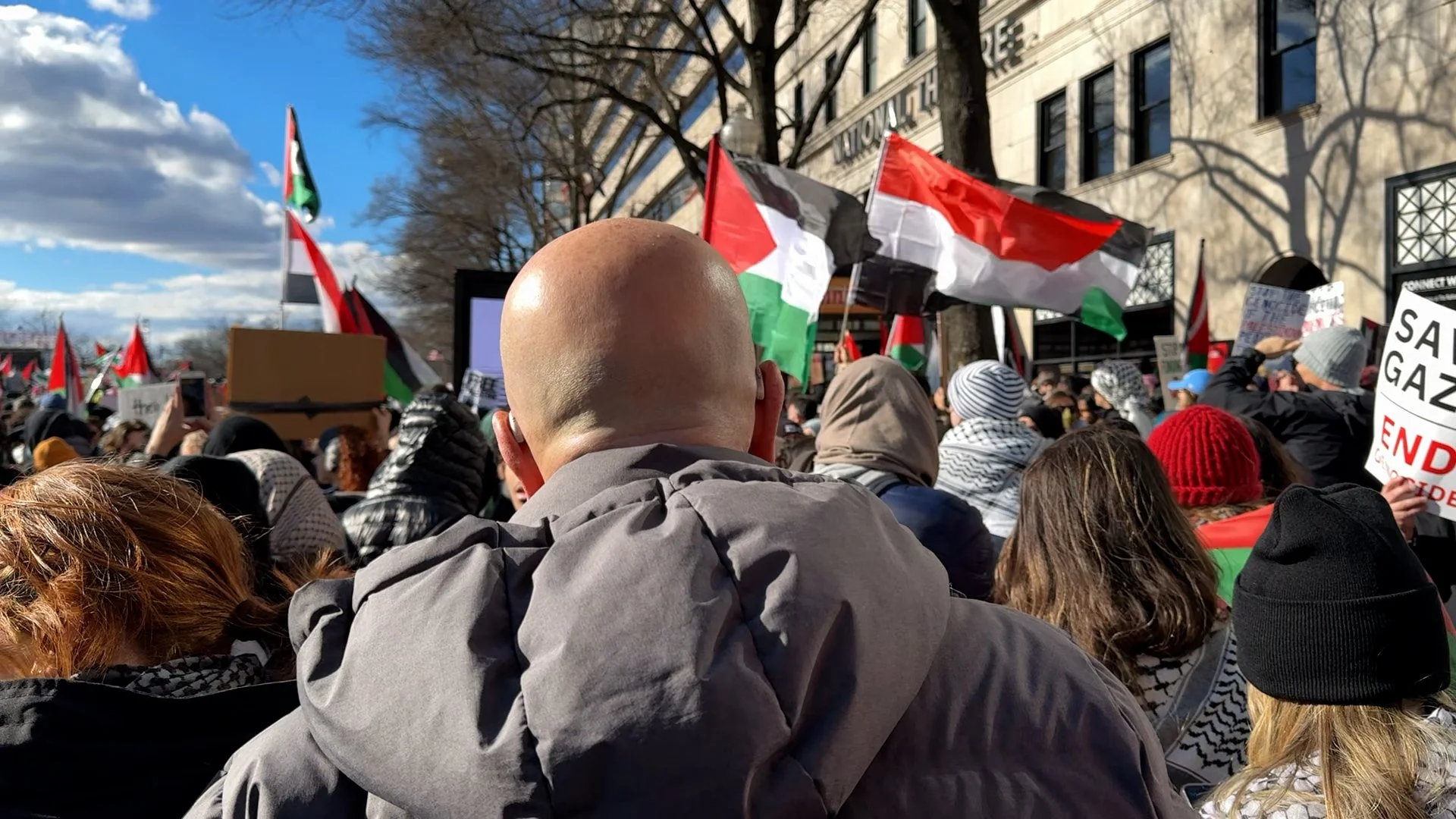 Crowd of people protesting with Palestinian flags and signs outside on a sunny day.