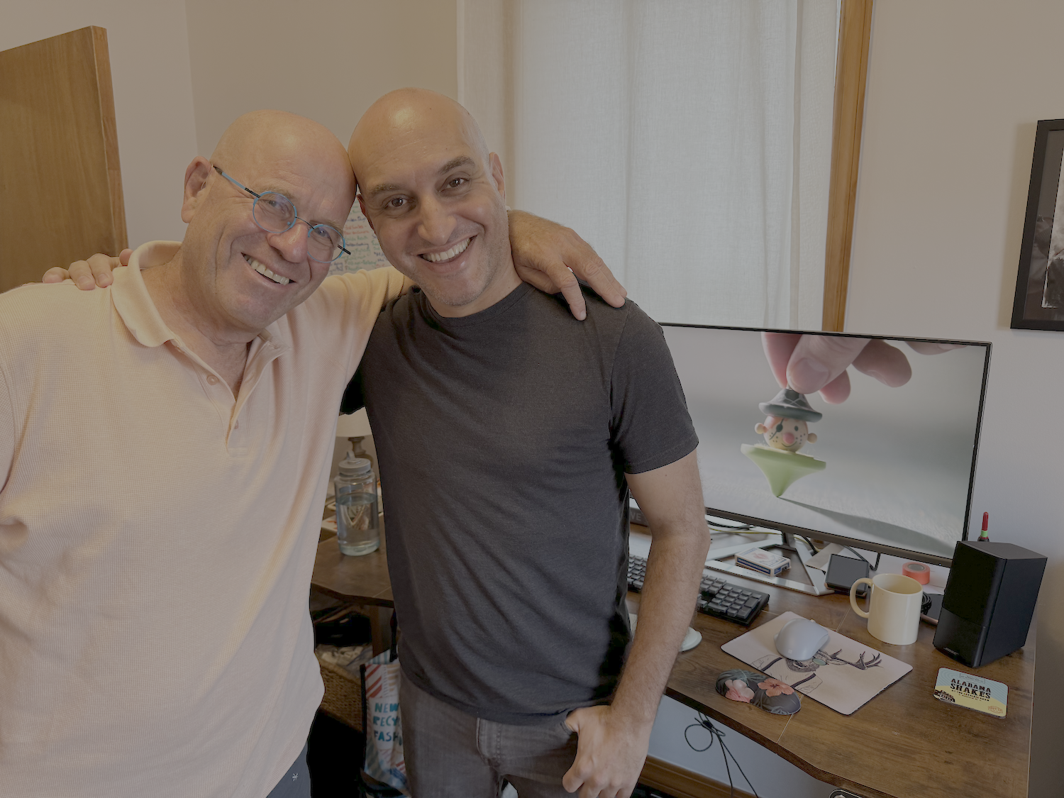 Two smiling men are standing close together, with one man having his arm around the other, in front of a desk with a computer and various items. A television screen behind them displays a close-up of a hand balancing a figure on a finger.