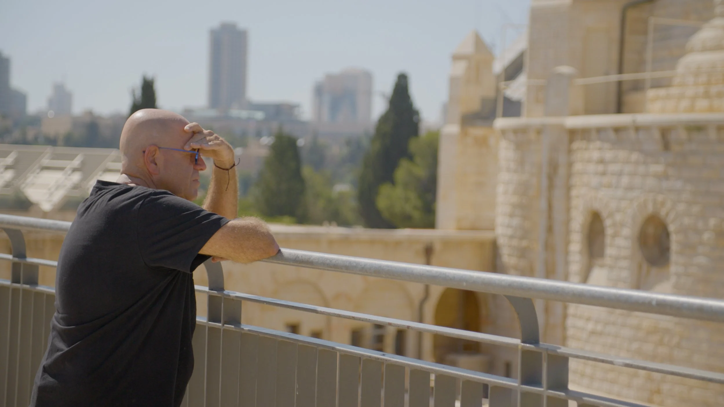 A man with a bald head, wearing glasses and a black T-shirt, leaning on a metal railing, looking out over a historic stone building with gothic architecture, with a city skyline in the background.