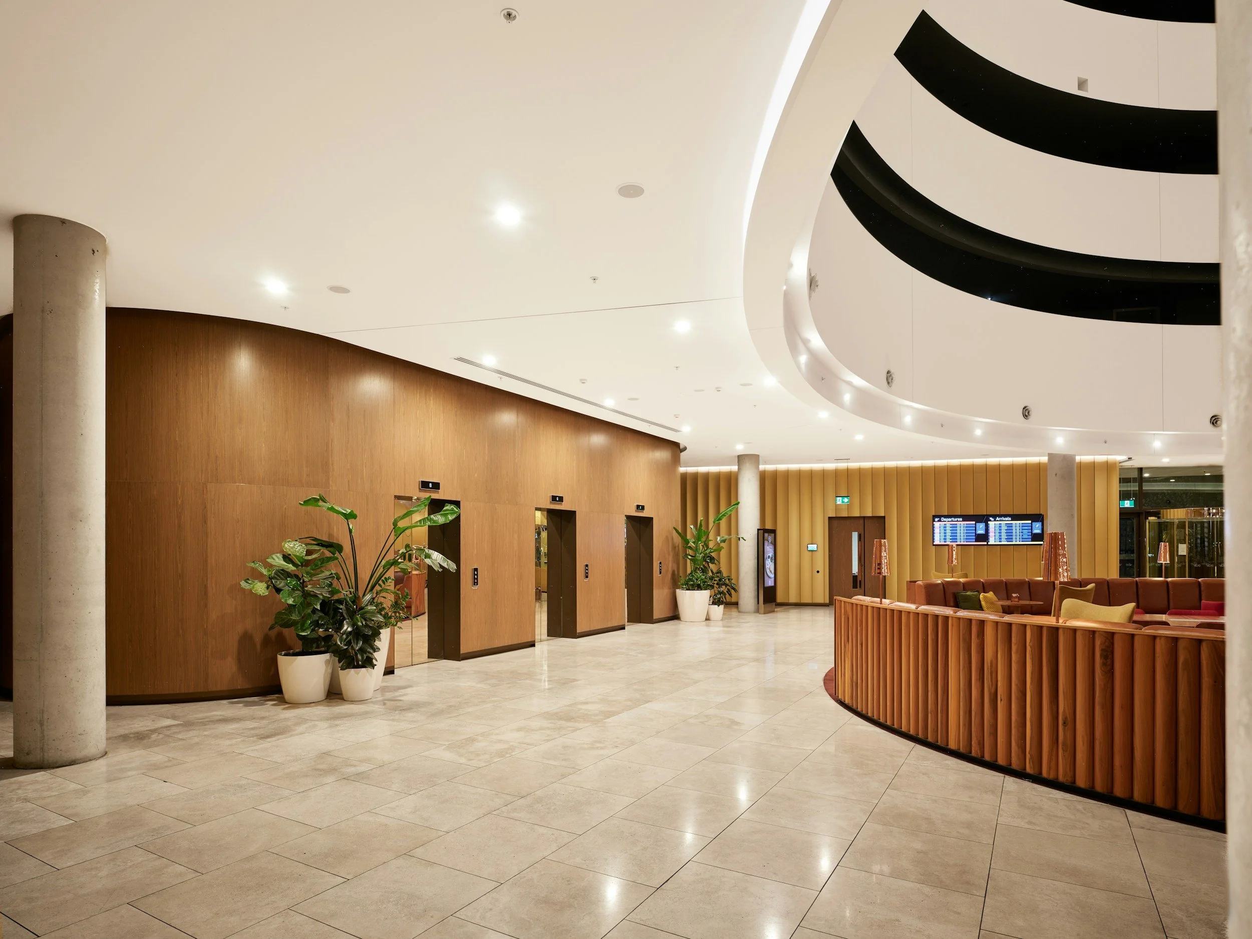 Interior of a modern airport lobby featuring three elevators on a wooden wall, potted plants, a seating area with brown and yellow cushions, and flight information screens on the wall.