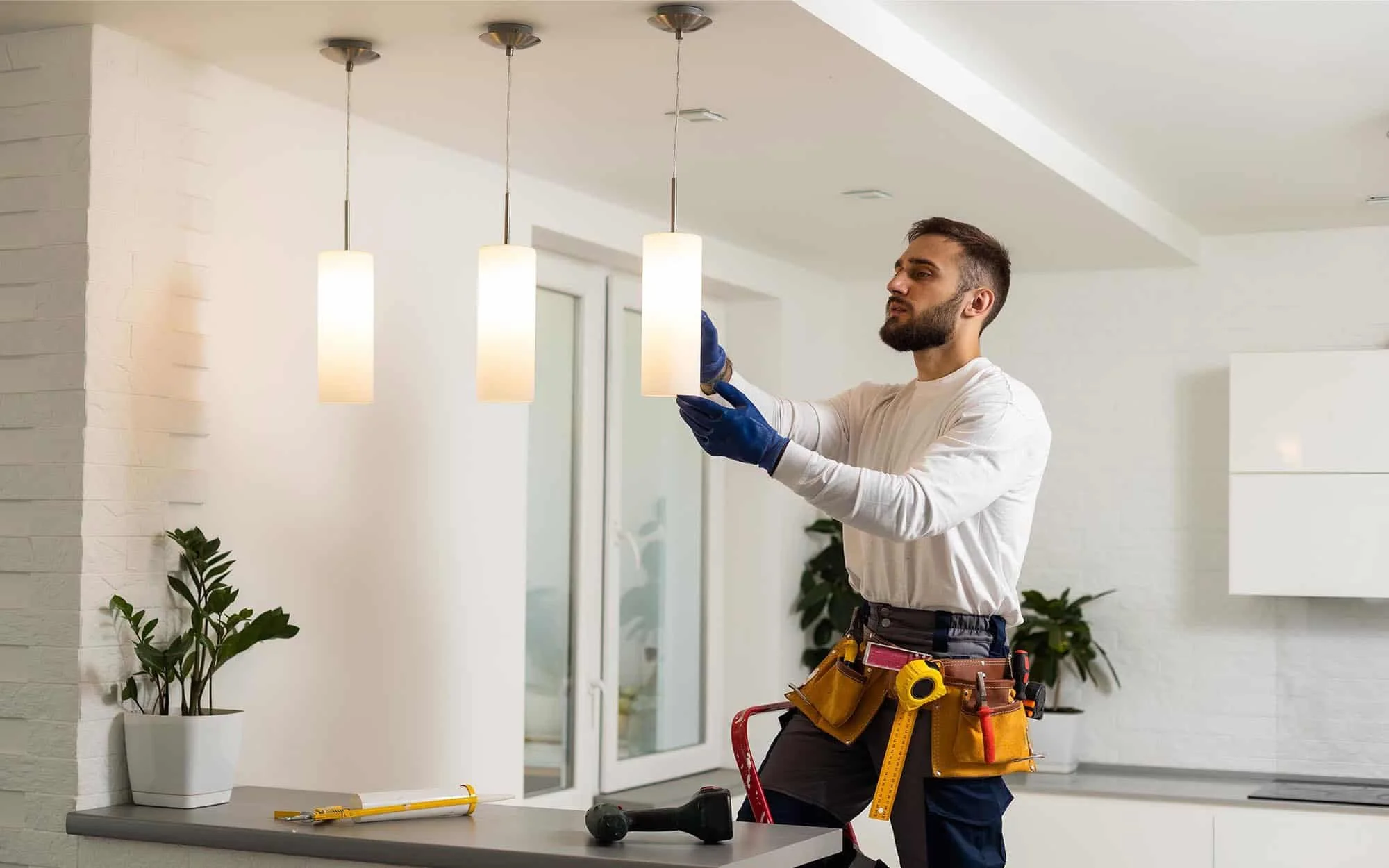 A man installing a ceiling light fixture in a modern kitchen, wearing work gloves and tool belt.