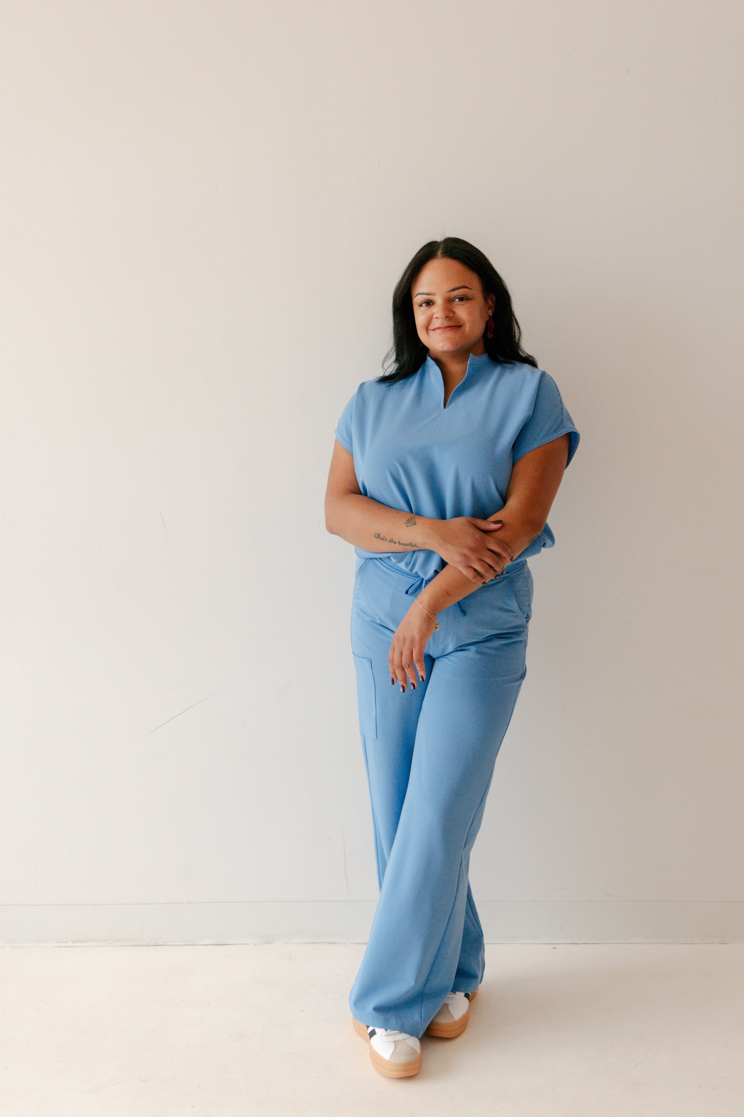 A woman with dark hair standing against a plain white wall, wearing a light blue medical uniform with rolled sleeves and wide-leg pants, paired with white sneakers, smiling and crossing her arms.