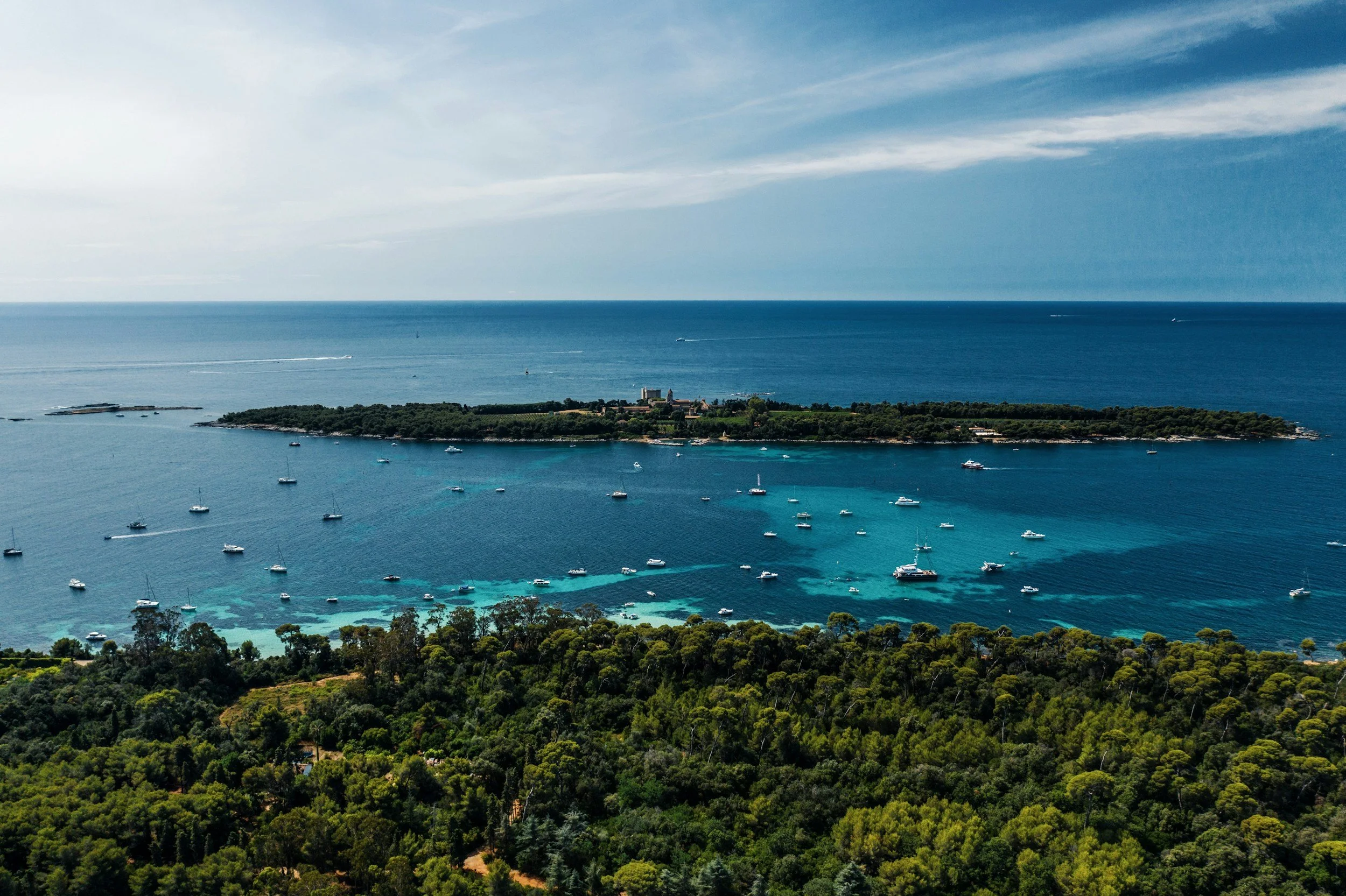 An aerial view of a coastal area with numerous sailboats and yachts anchored in the turquoise and blue water, a forested shoreline, and a distant horizon with a mix of blue sky and scattered clouds.