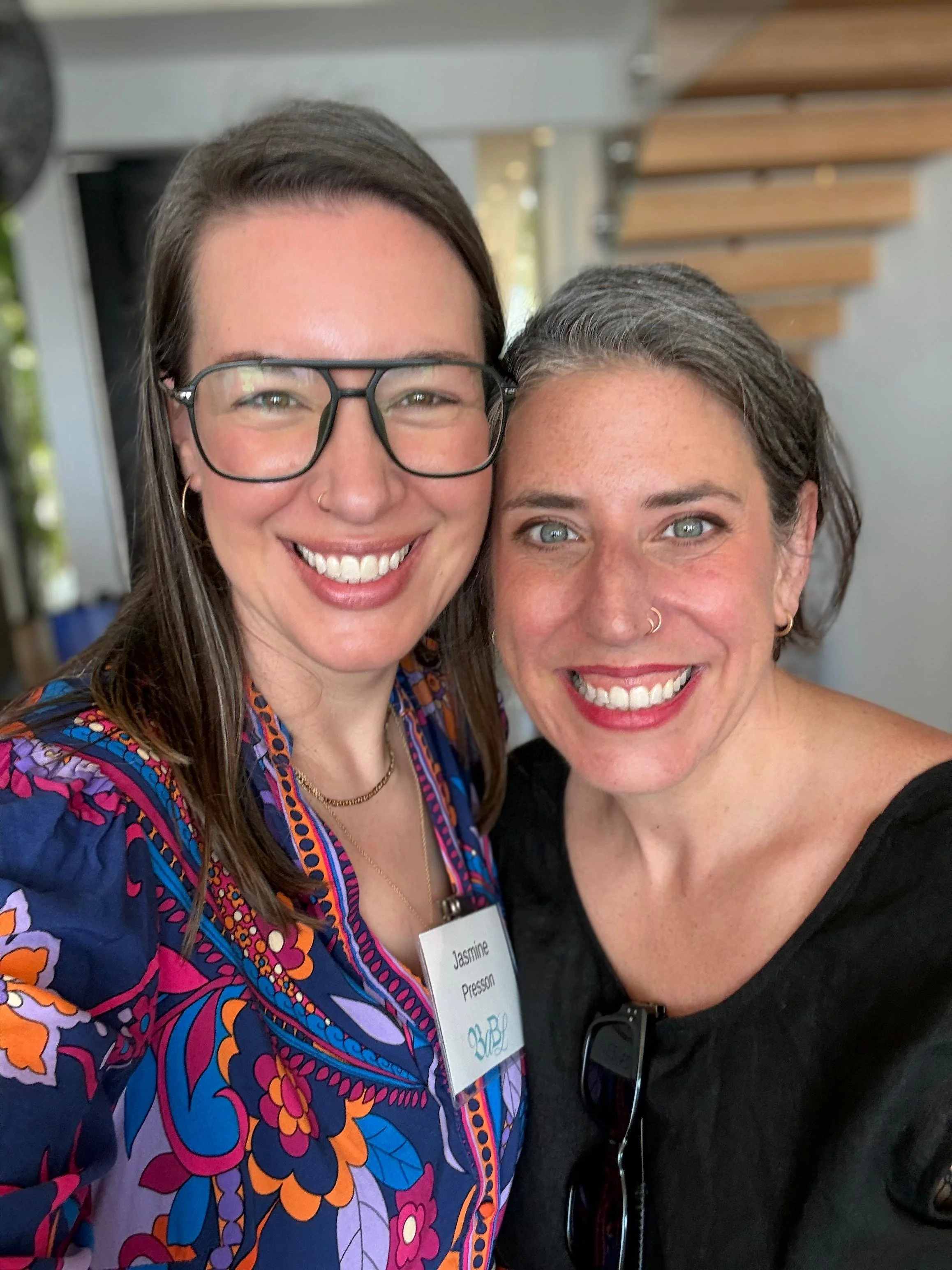 Two women smiling and posing for a selfie together. One woman has long brown hair, glasses, and is wearing a colorful, patterned shirt. The other woman has short gray hair, a nose ring, and is wearing a black top.