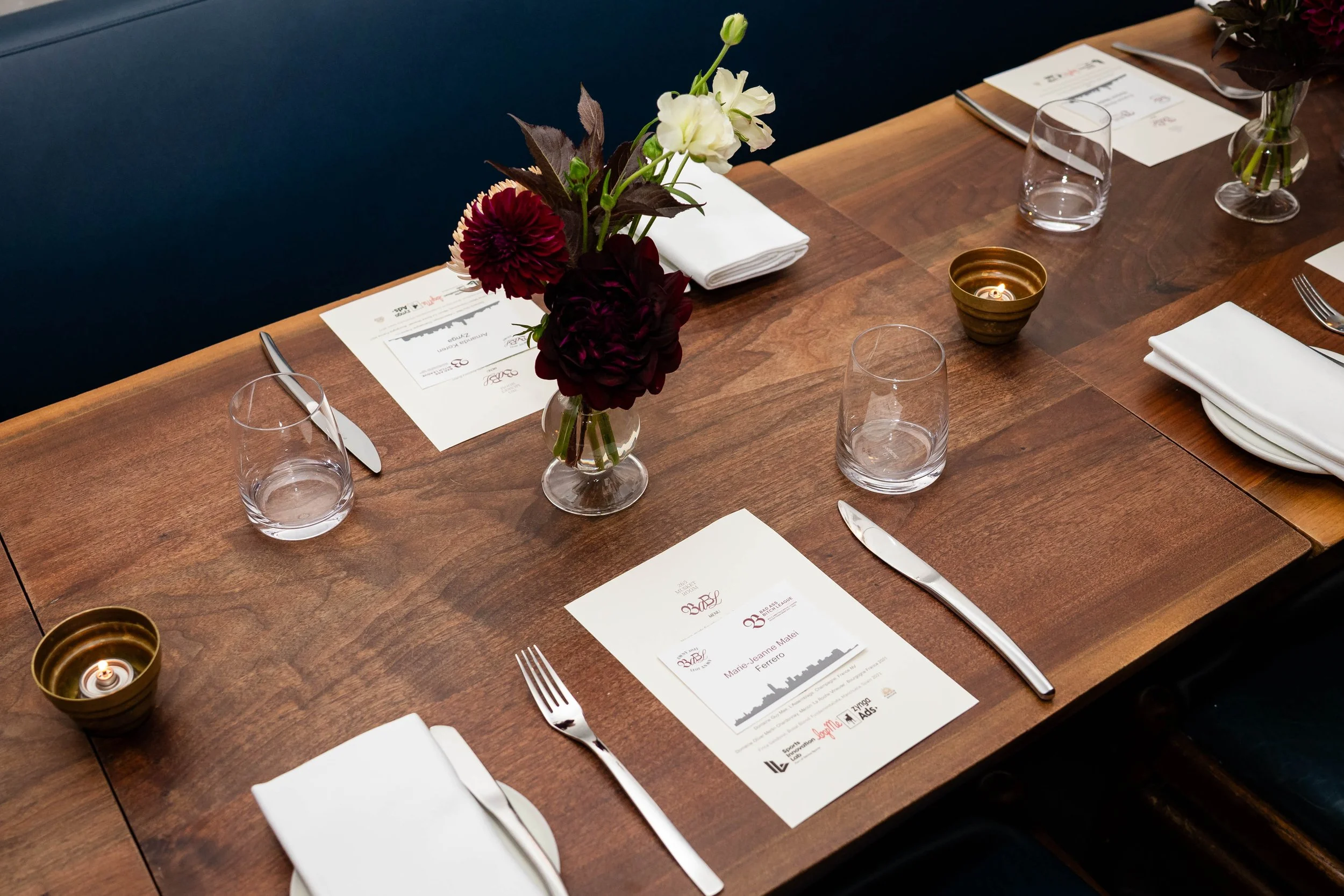 A dining table set with white plates, silverware, drinking glasses, white napkins, a small flower arrangement with white and dark red flowers, and a lit candle in a small brass holder.