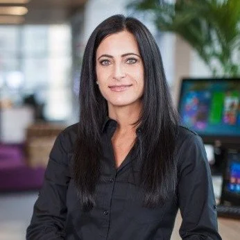 A woman with long black hair, wearing a black shirt, sitting indoors with a computer monitor in the background.
