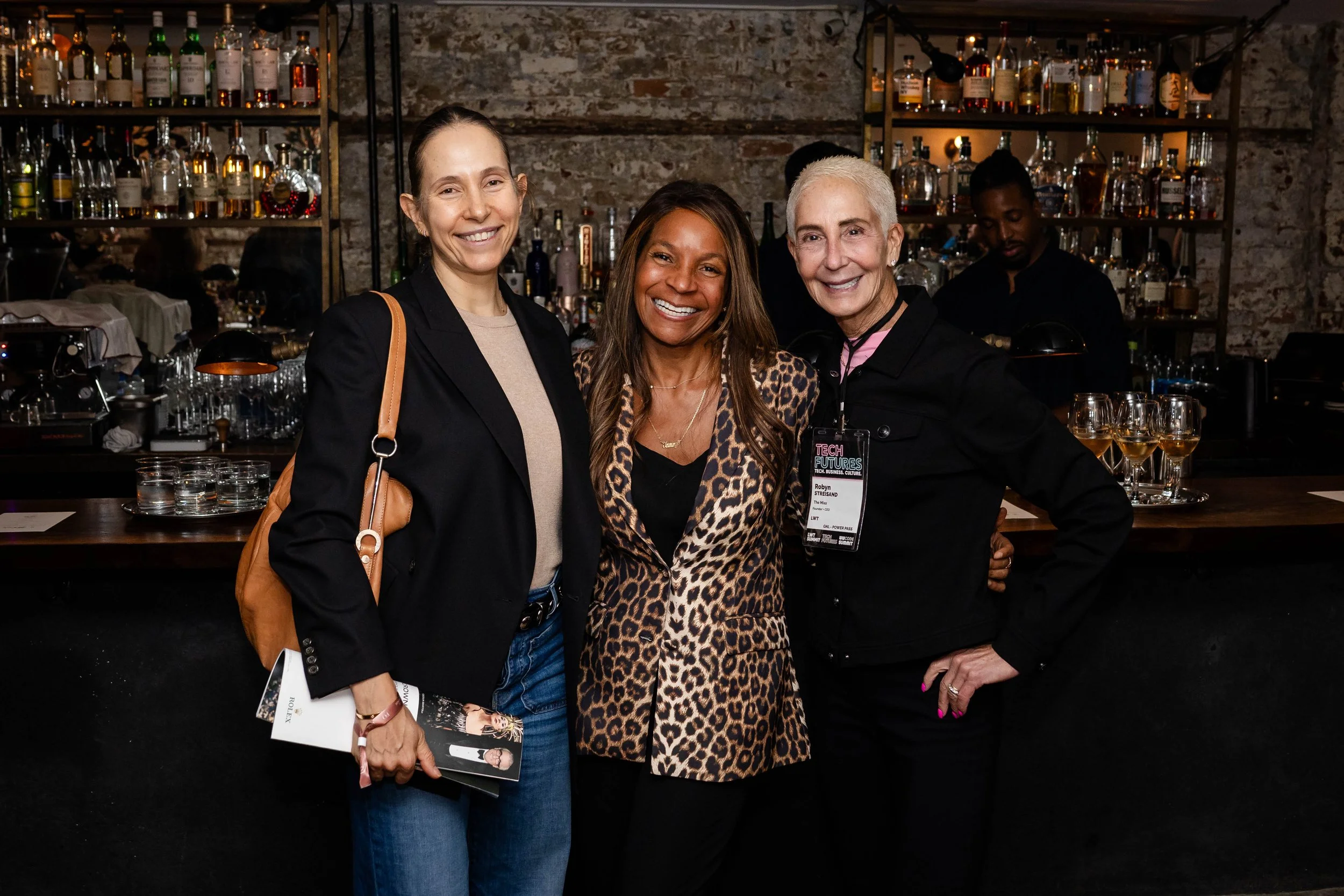 Three women and one man smiling for a photo in a bar with exposed brick walls and shelves of bottles in the background. The woman in the middle wears a leopard print coat, the woman on the left wears a black blazer and beige shirt, and the woman on the right wears black with a name tag that reads 'Robyn'.