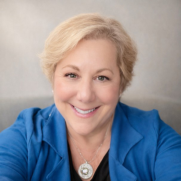 A smiling middle-aged woman with short blonde hair, wearing a blue blazer and a silver necklace, against a plain background.