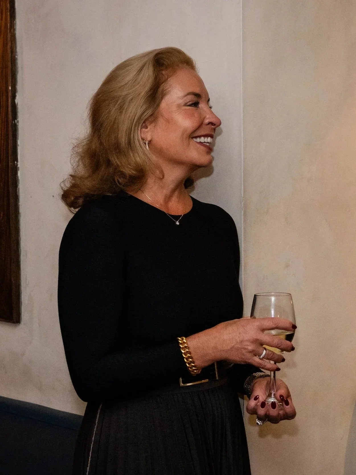 A woman with blonde hair wearing a black top and gold jewelry holding a glass of white wine, smiling and standing against a plain wall.