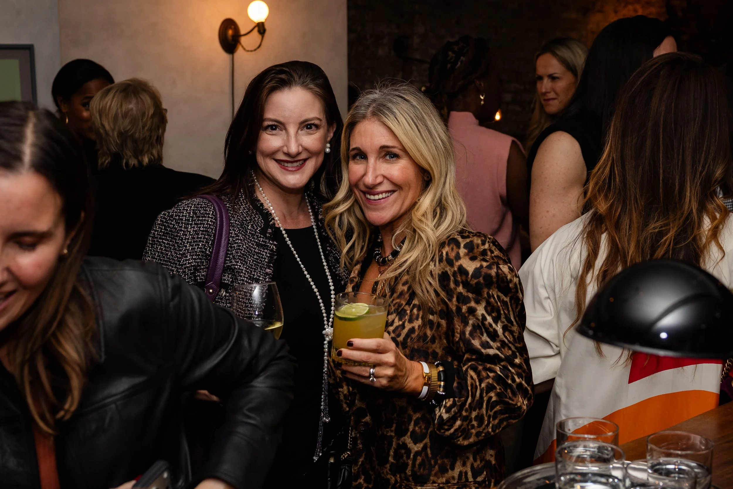 A group of women at a social gathering, with two women smiling and holding drinks in the foreground. One woman has dark hair and is wearing pearls, the other has blonde hair and is dressed in a leopard print top. The background shows other women engaged in conversation, with warm lighting and dark brick walls.