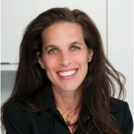 A smiling woman with long dark hair, wearing a black blazer, standing in front of a light-colored background.