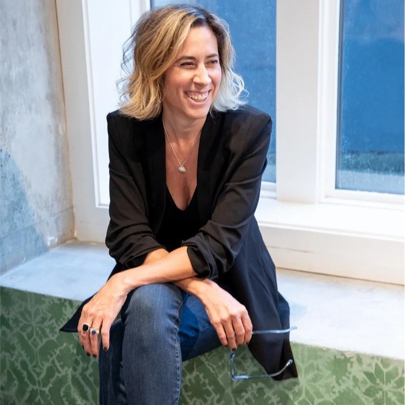 A woman with blonde, wavy hair, wearing a black blazer and jeans, smiling and sitting on a windowsill.
