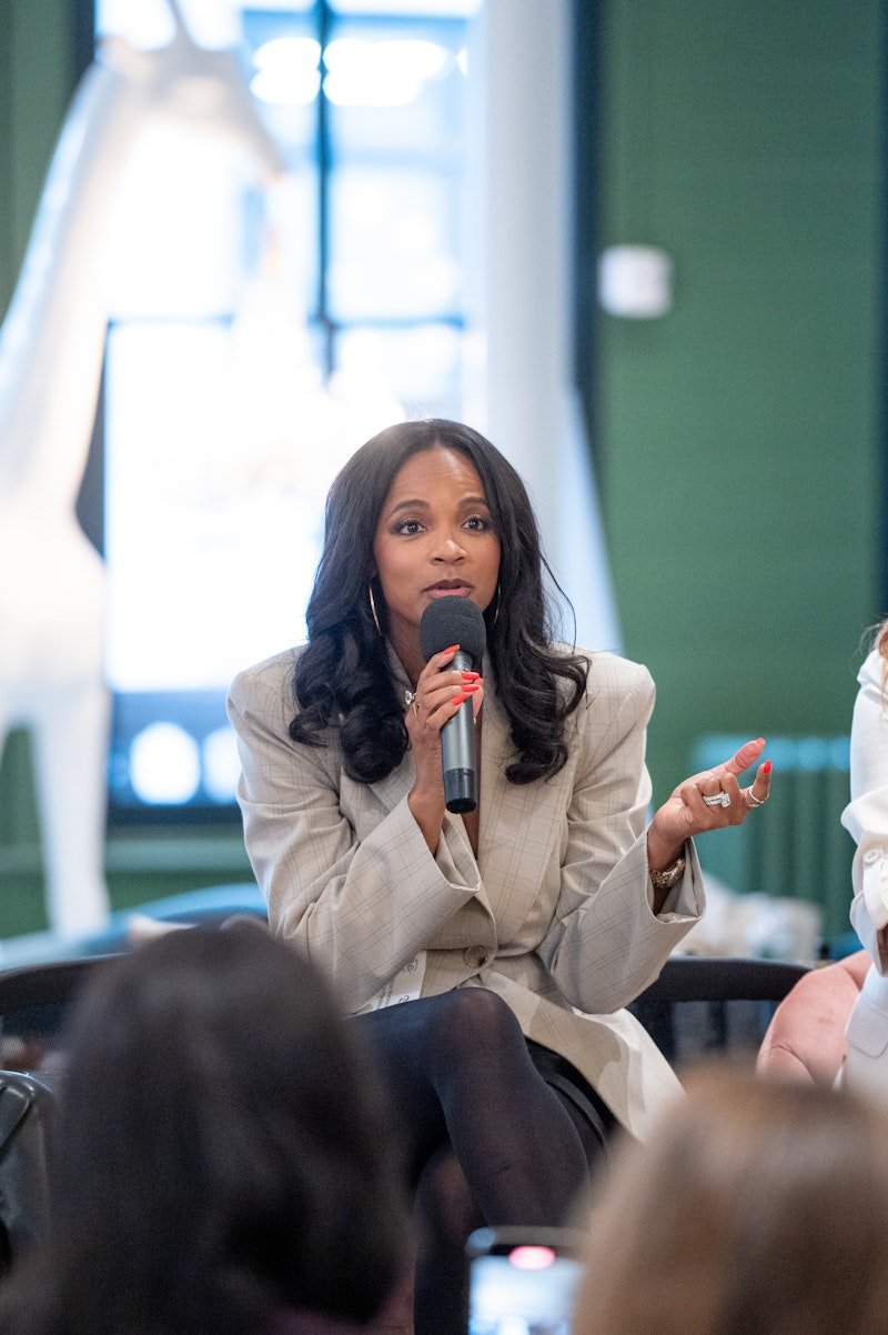 A woman with long dark hair, wearing a beige blazer and black tights, is sitting and speaking into a microphone during a panel discussion or interview.