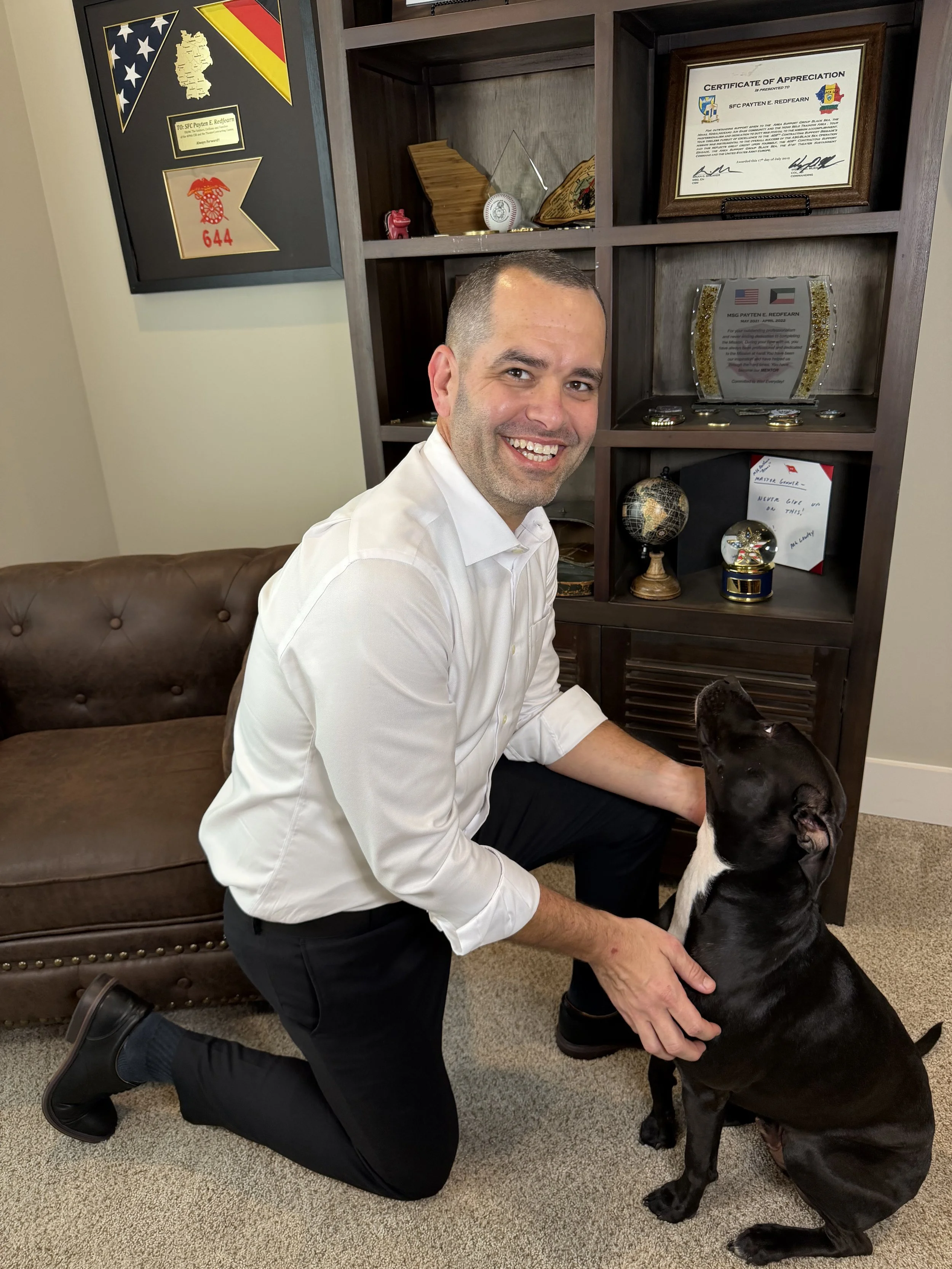 A man kneeling on one knee, smiling, with a black and white dog that is sitting and looking up at him. They are indoors in front of a wooden shelf with awards and framed certificates, with a leather sofa to the left.