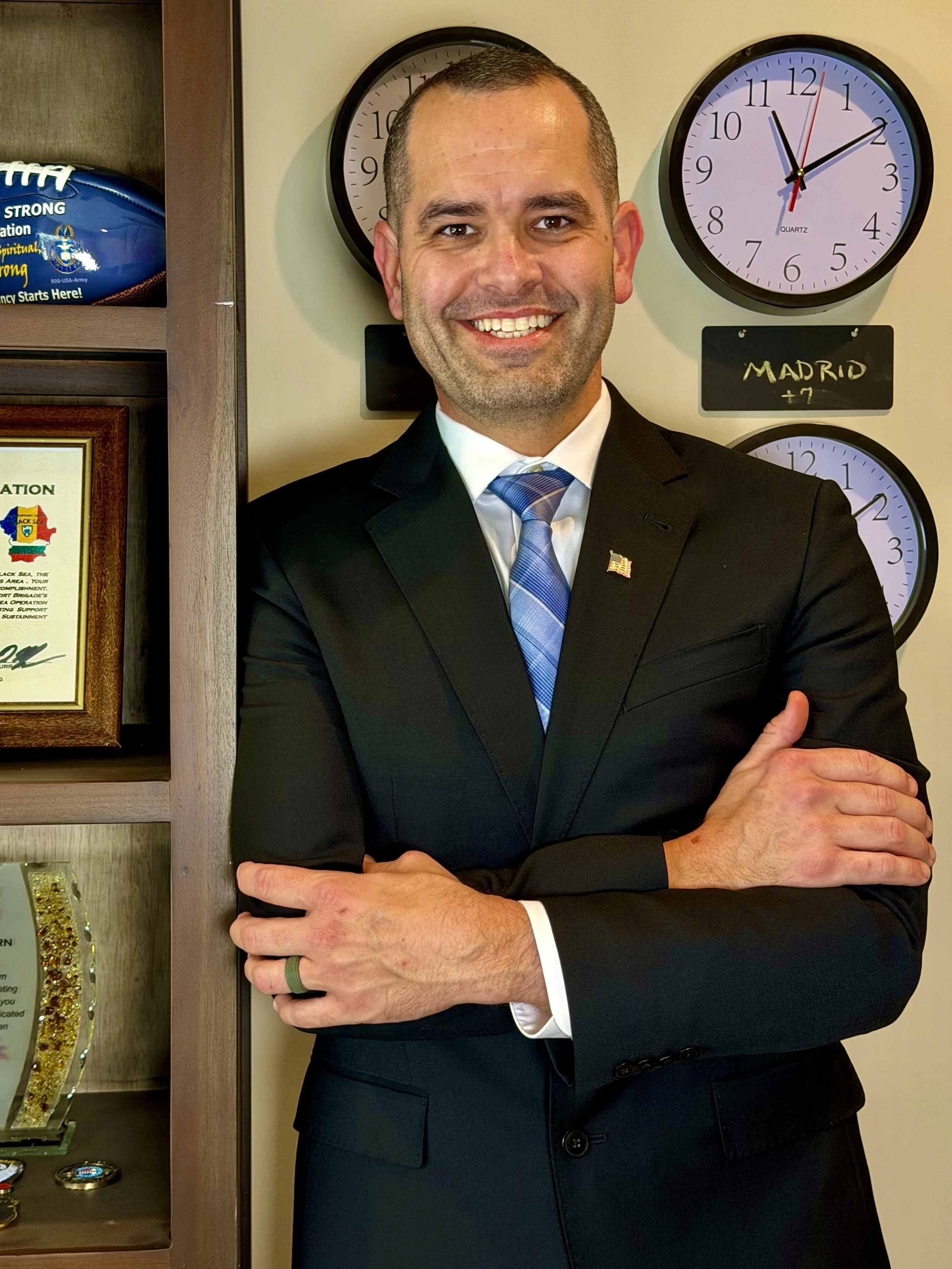 A man dressed in a black suit and blue tie standing in front of three clocks on a beige wall, with a framed certificate and trophies on a shelf next to him.