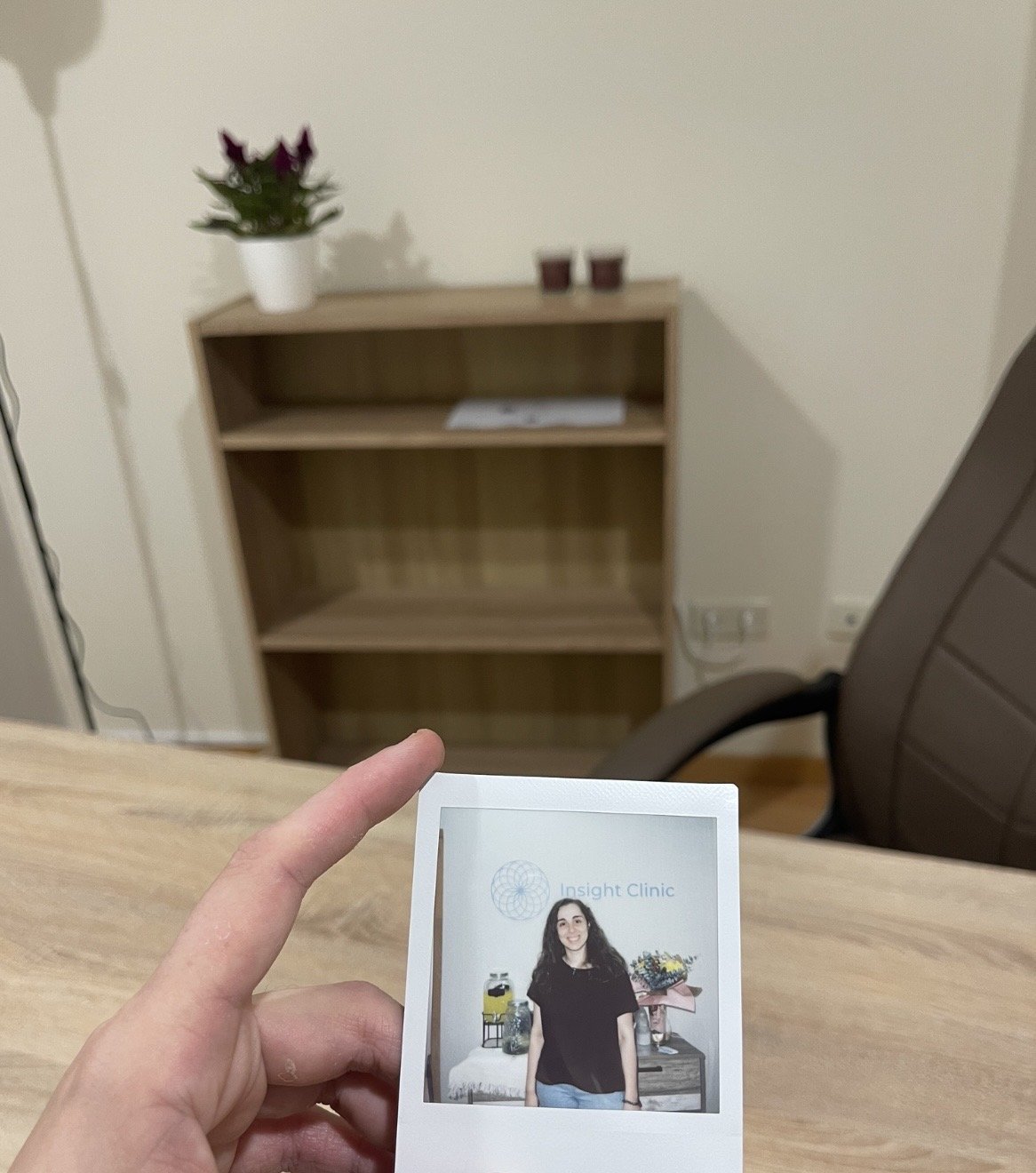 Gabinete de psicologia, psicóloga segurando uma foto impressa de uma mulher sorridente, com fundo de uma sala com móveis e decoração.