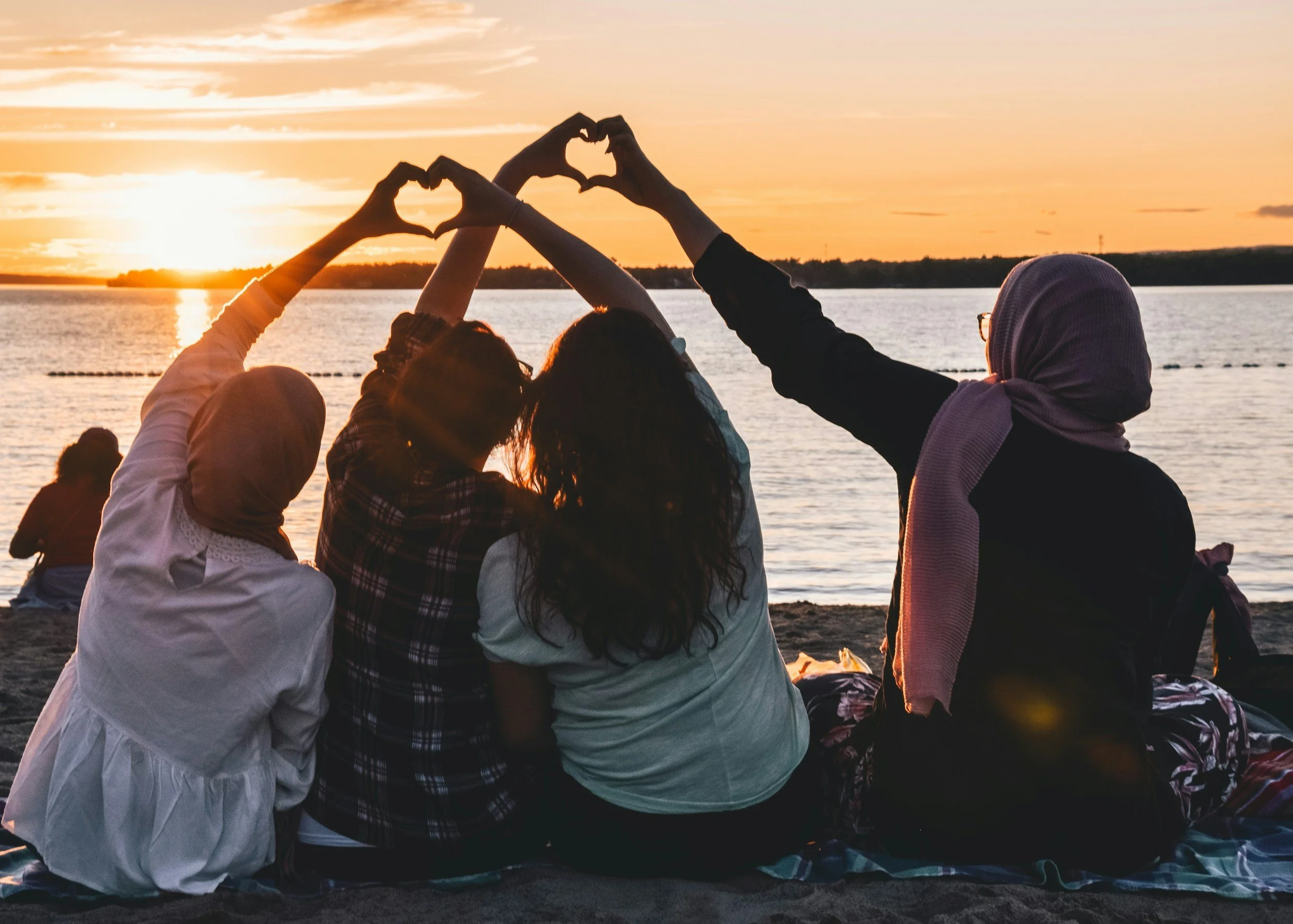 Group of five people sitting on a beach during sunset, making a heart shape with their hands, with water and the setting sun in the background.