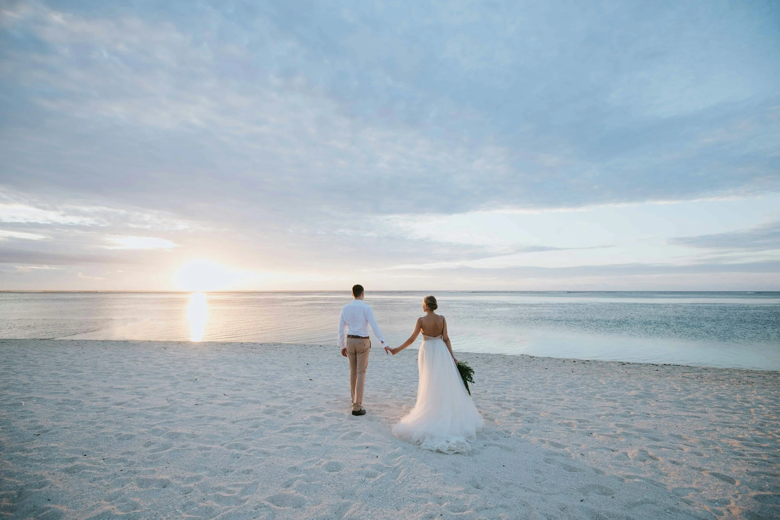 A couple in wedding attire holding hands on a sandy beach during sunset, overlooking the ocean.