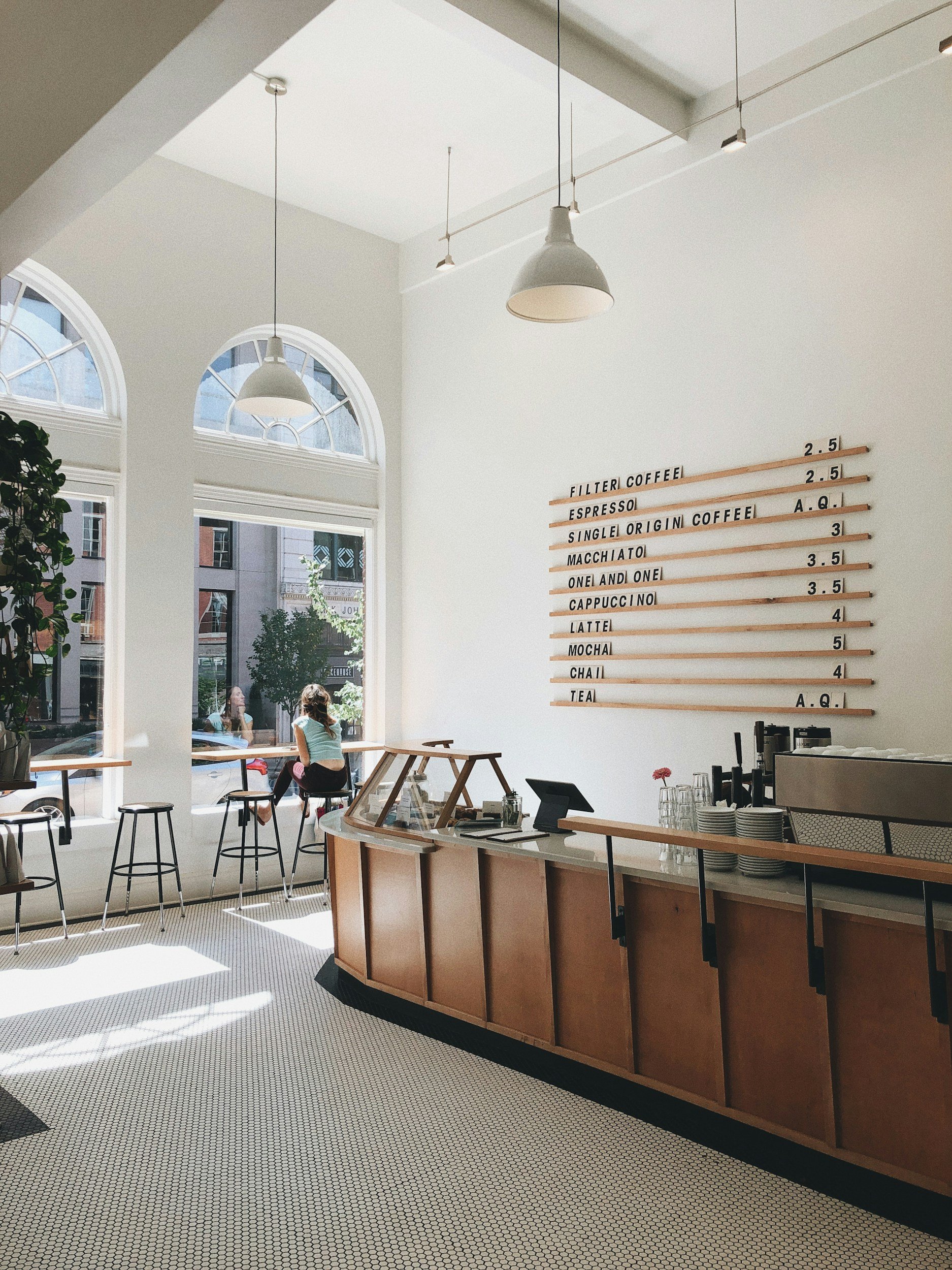 Interior of a modern coffee shop with a white counter, menu board, and large arched windows allowing sunlight in. Several hanging pendant lights are visible, and a customer is sitting by the window.