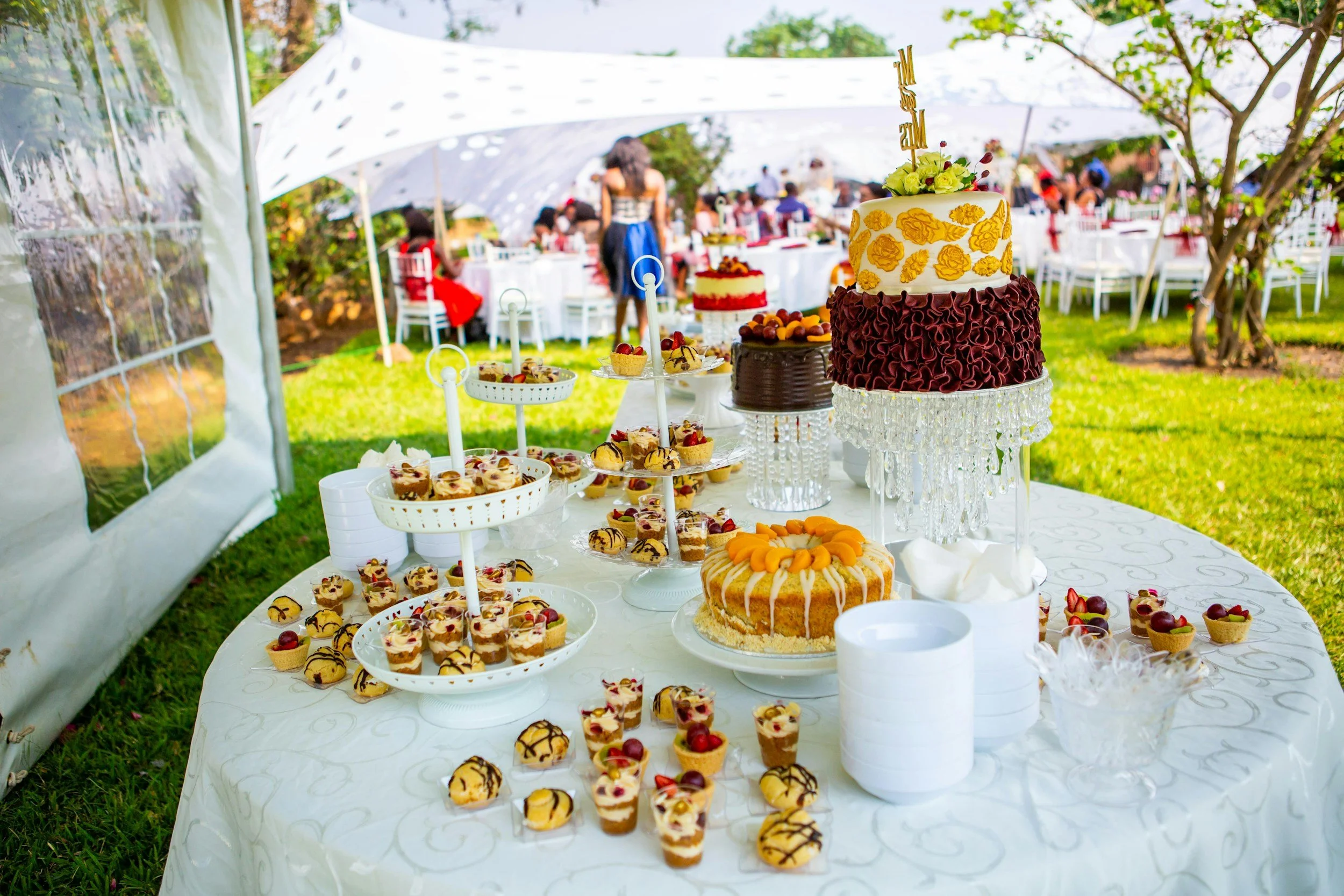 Decorated table with cakes and desserts at an outdoor celebration under a white tent with people in the background.