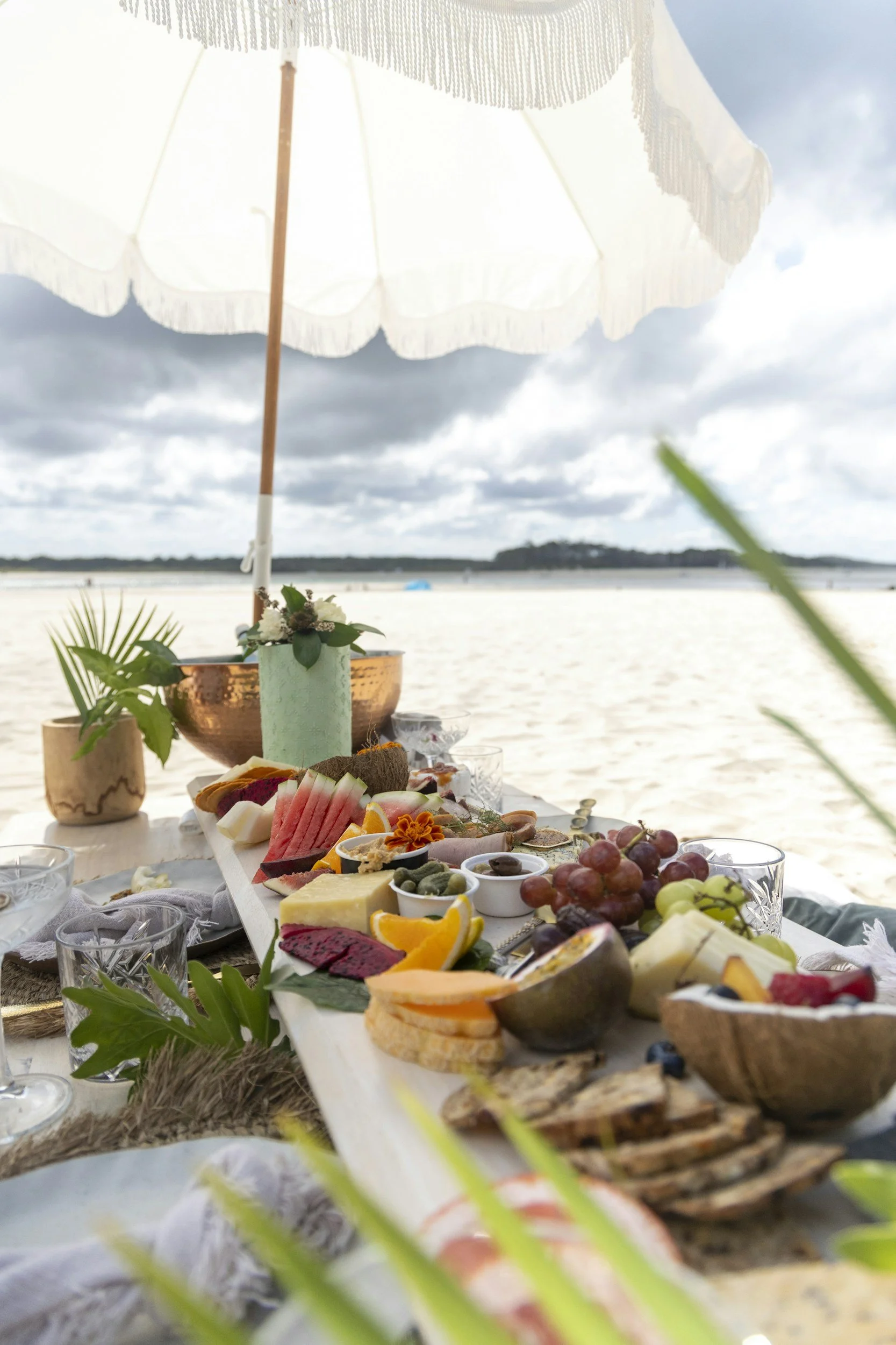 A beachside picnic with a table filled with a variety of cheese, fruits, and snacks, under a white parasol on sandy beach with cloudy sky.