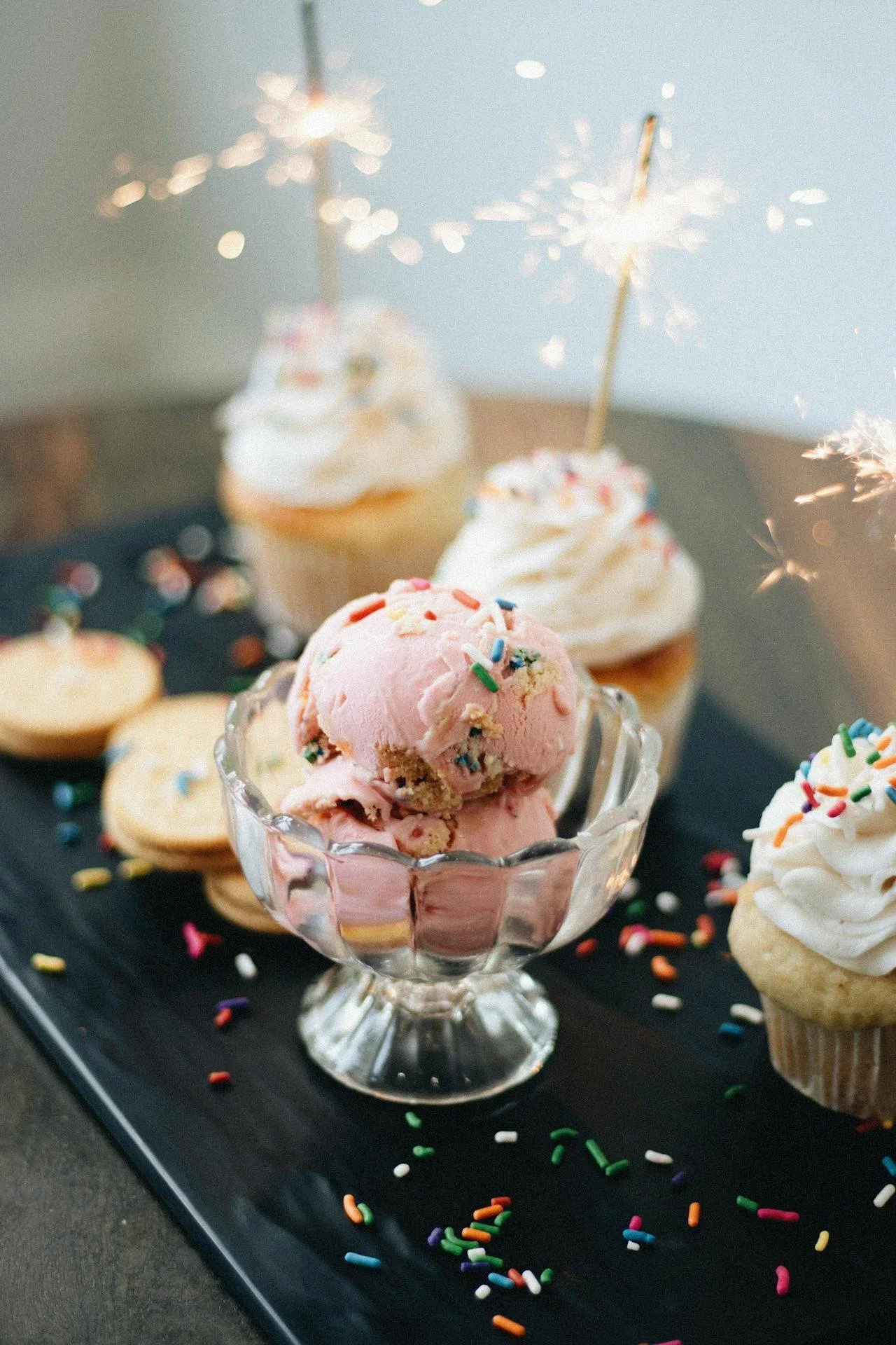 Cupcakes with white frosting and colorful sprinkles, decorated with sparklers, and a bowl of pink ice cream with sprinkles on a black tray with scattered sprinkles.