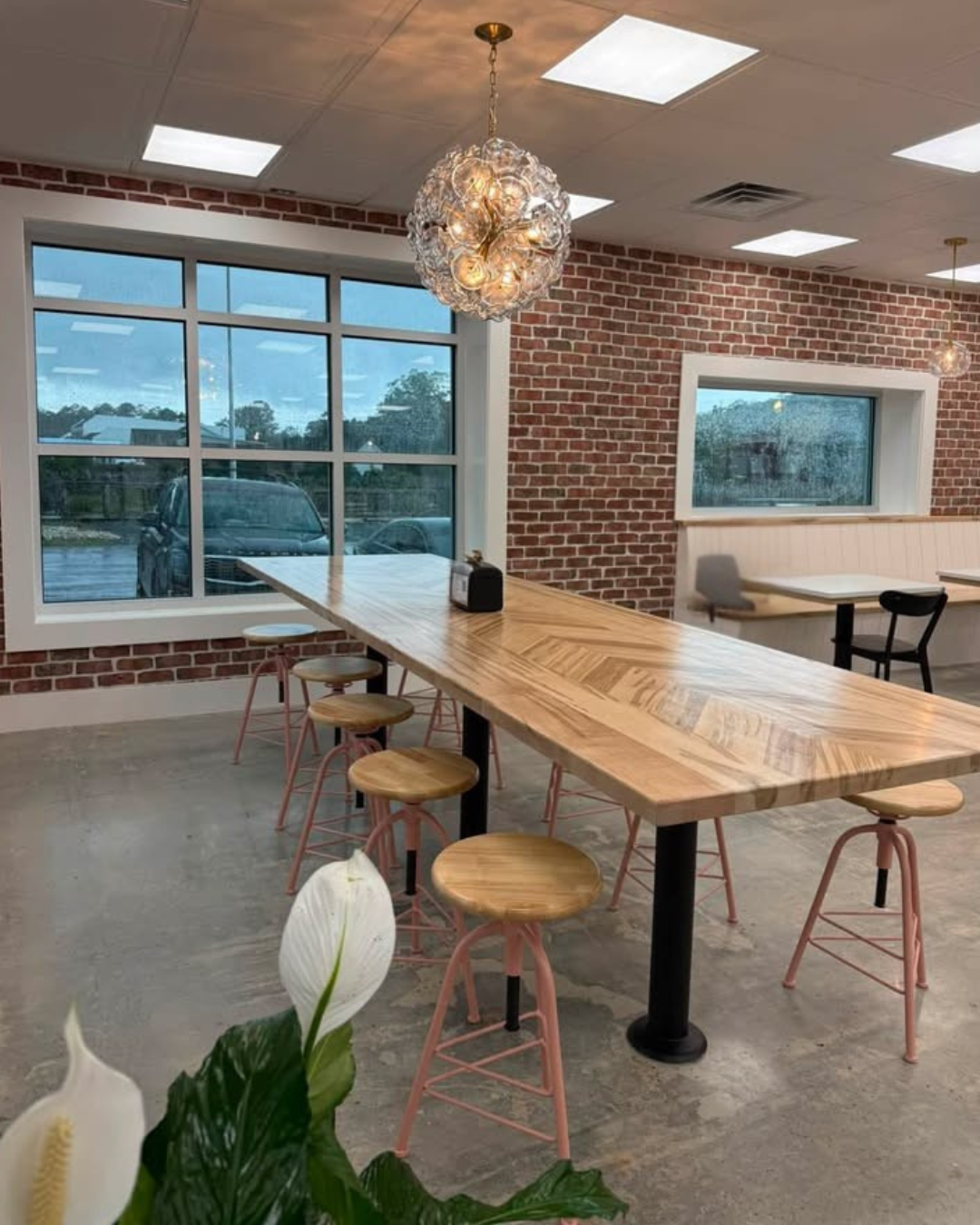 The inside of Lilly Bird's Creamery: A modern cafe interior with a large wooden table, pink barstools, brick walls, large window, and decorative hanging light fixture.