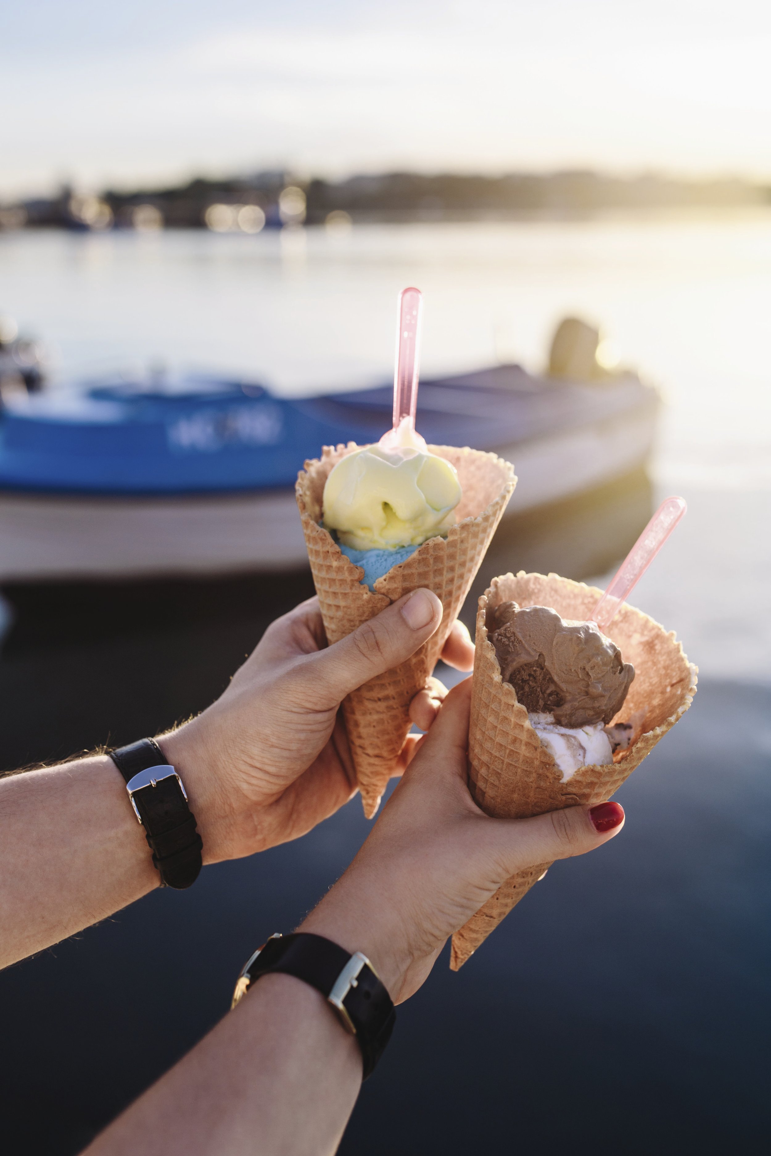 Two hands holding ice cream cones with a boat and water in the background during sunset.