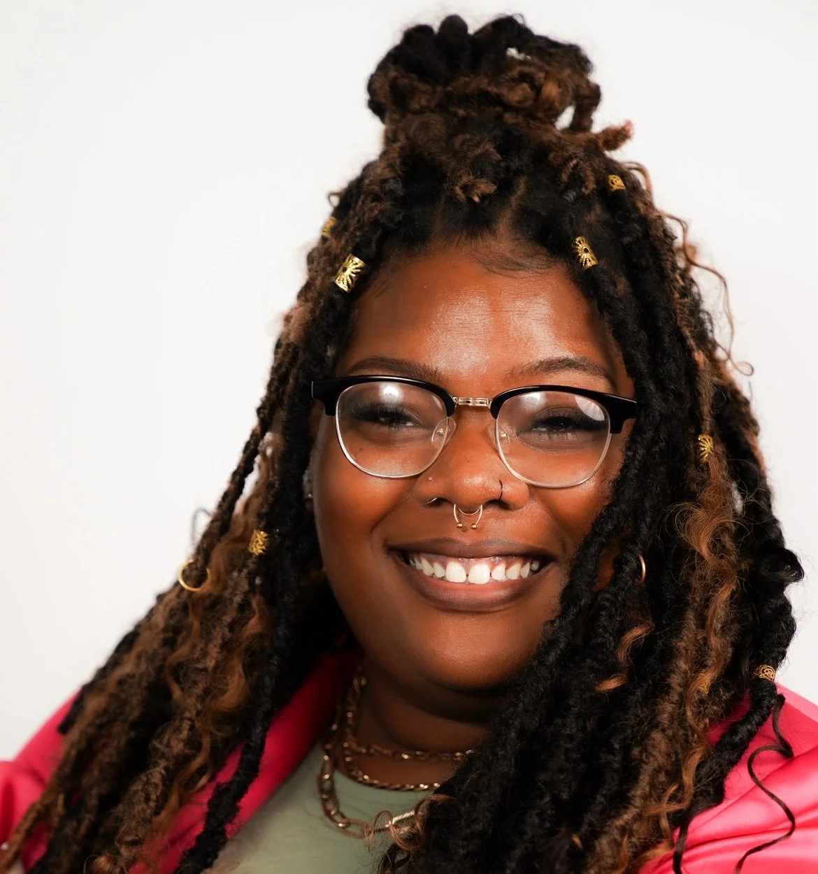 A smiling woman with glasses and dreadlocks adorned with gold cuffs, wearing a pink jacket over a light green top, against a plain white background.