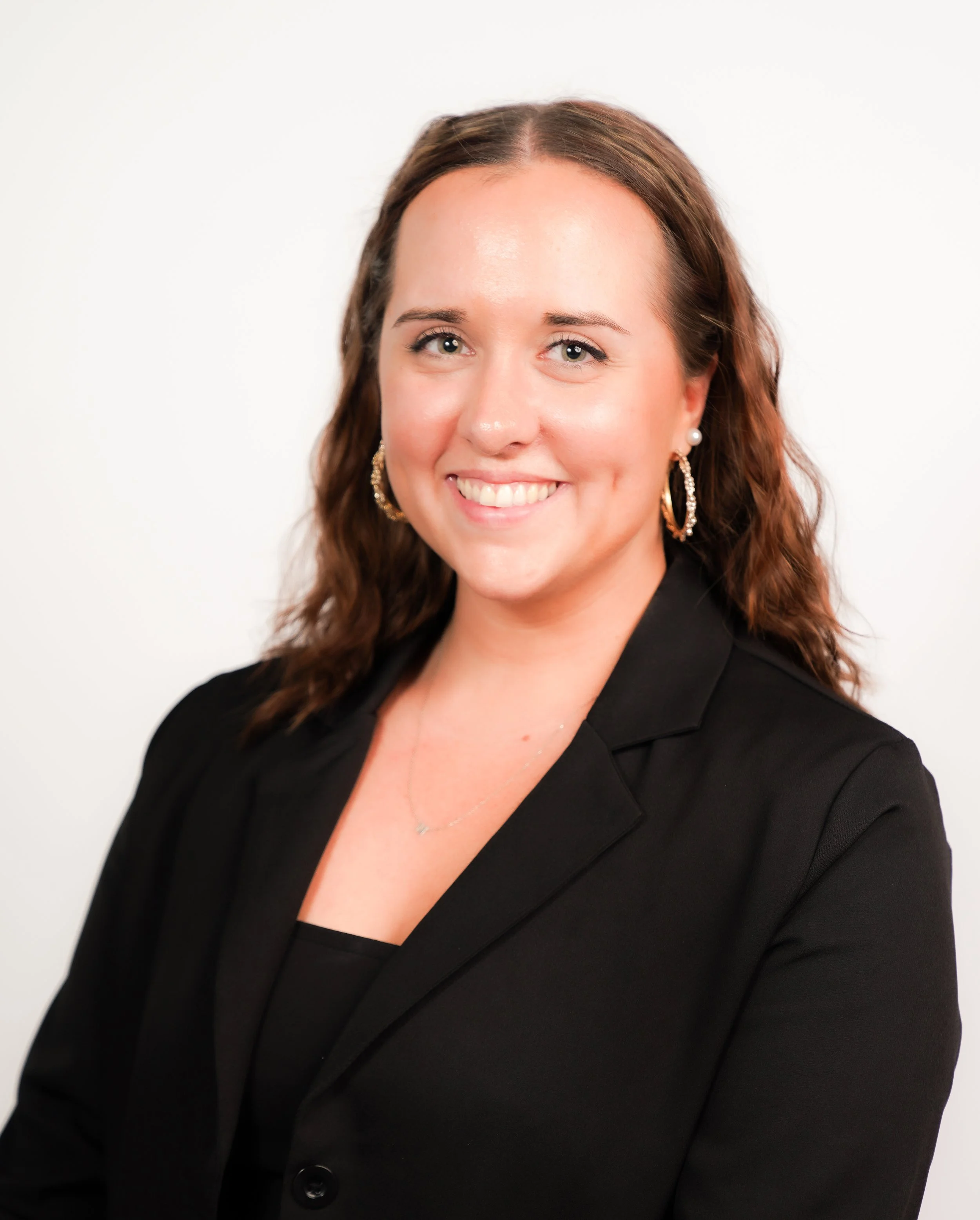 A woman with shoulder-length brown hair, smiling, wearing a black blazer, gold earrings, a pearl earring, and a delicate necklace, against a plain white background.