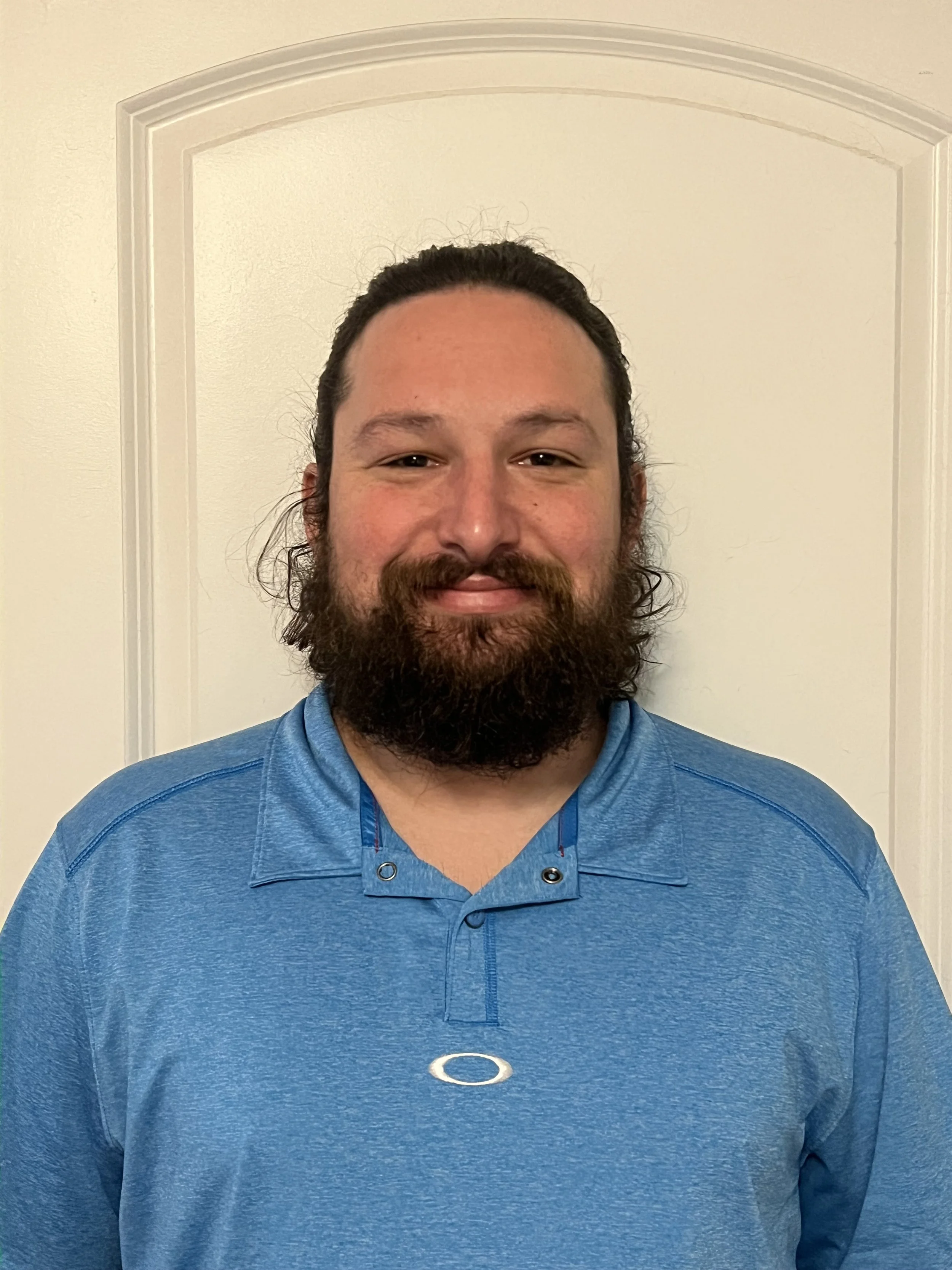 A man with a beard and long dark hair smiling, wearing a blue collared athletic shirt, standing in front of a white door.