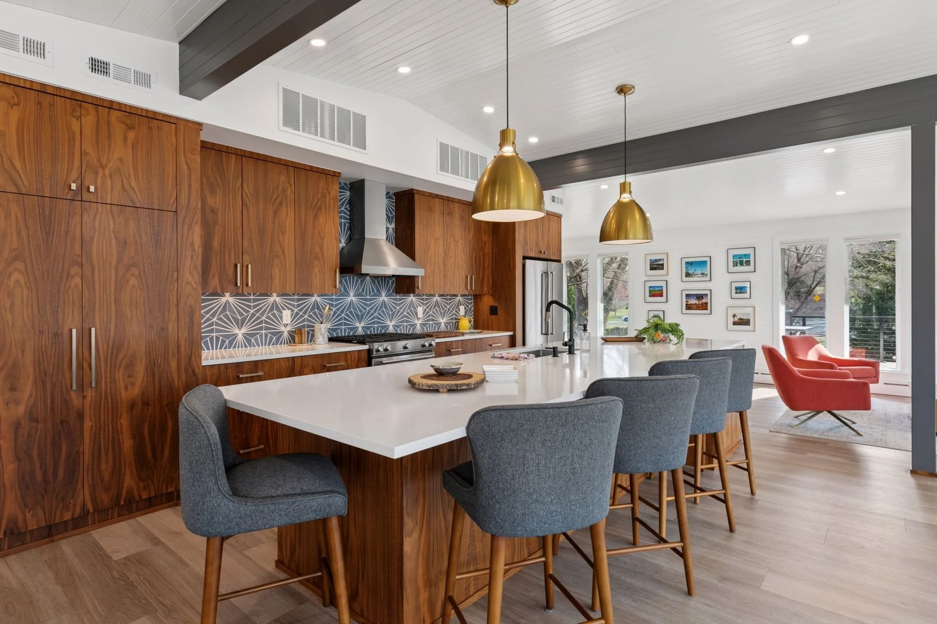 Modern kitchen with wooden cabinets, white island, gold pendant lights, and a dining area with red and gray chairs, large windows, and artwork on the wall.