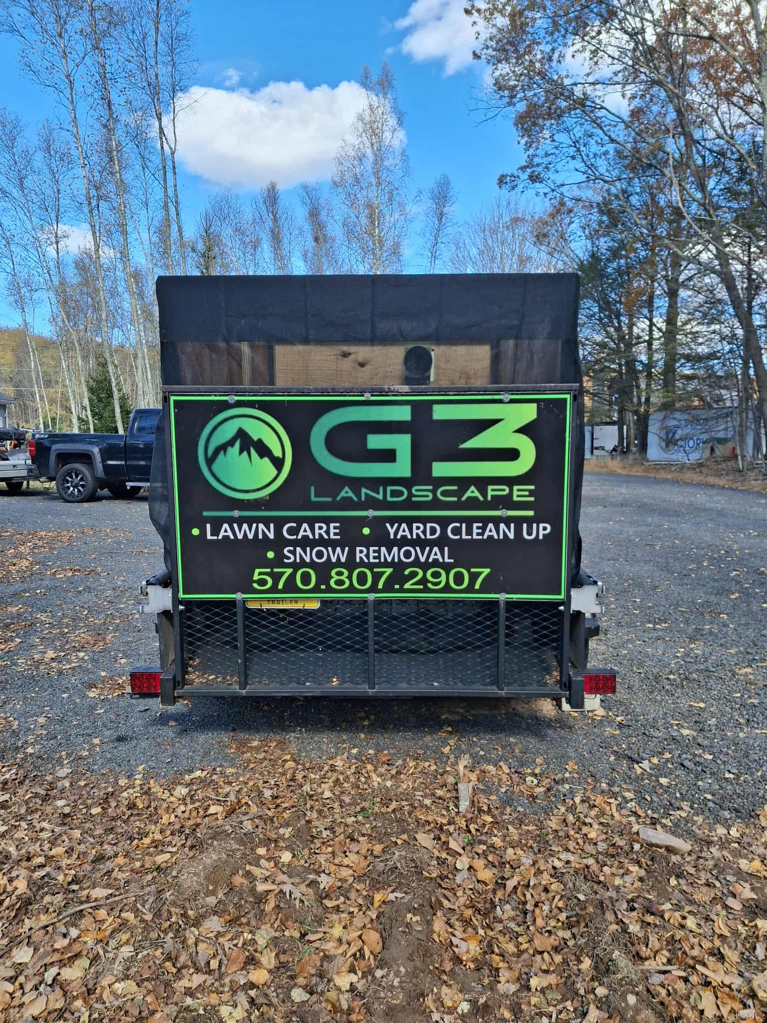 Trailer with G3 Landscape sign advertising lawn care, yard clean up, snow removal, and contact phone number 570-807-2907, in an outdoor area with trees and parked vehicles.